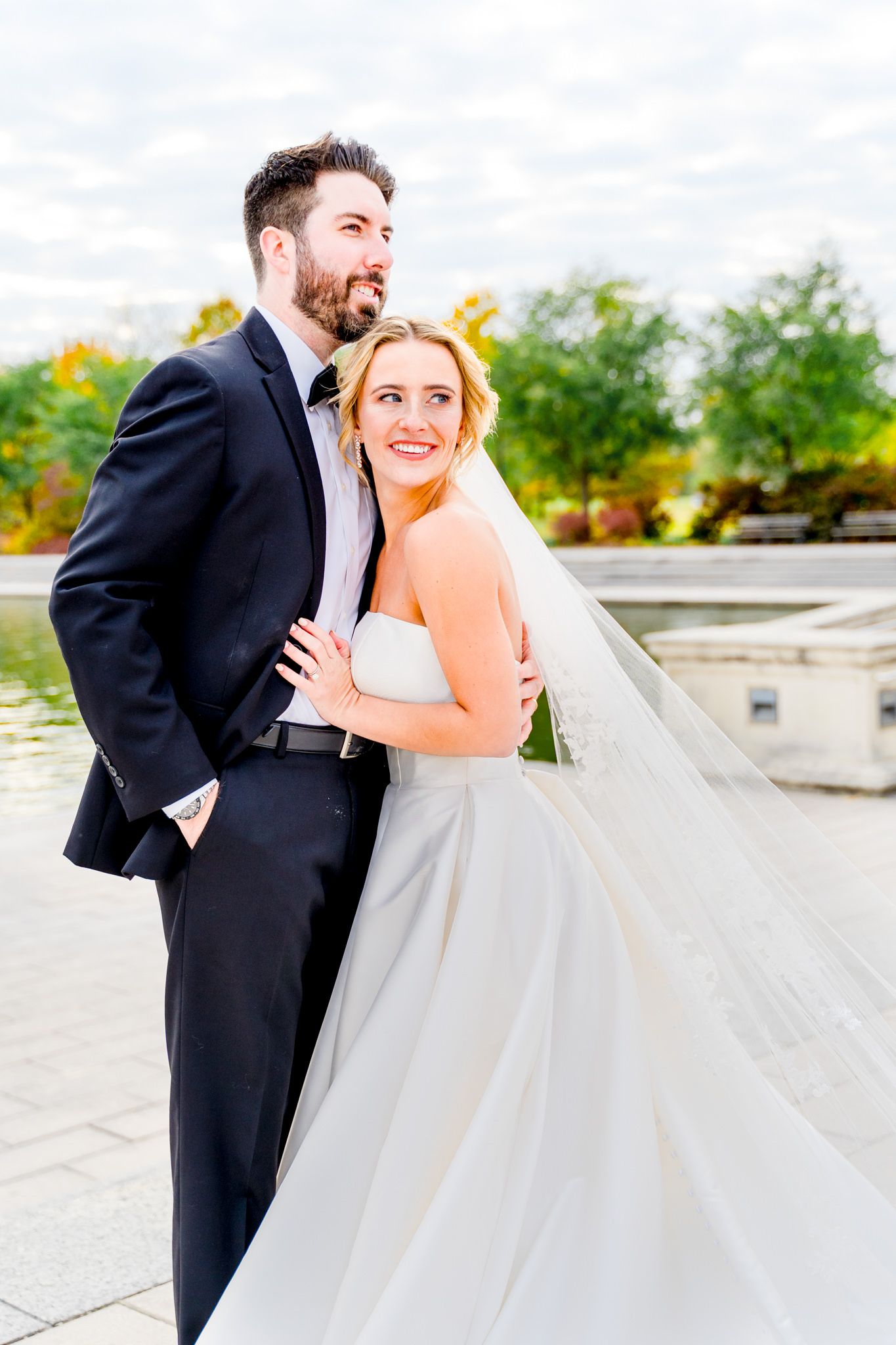 bride's wedding dress and veil fanned out next to groom under blue sky with clouds for Forest Park STL fall wedding