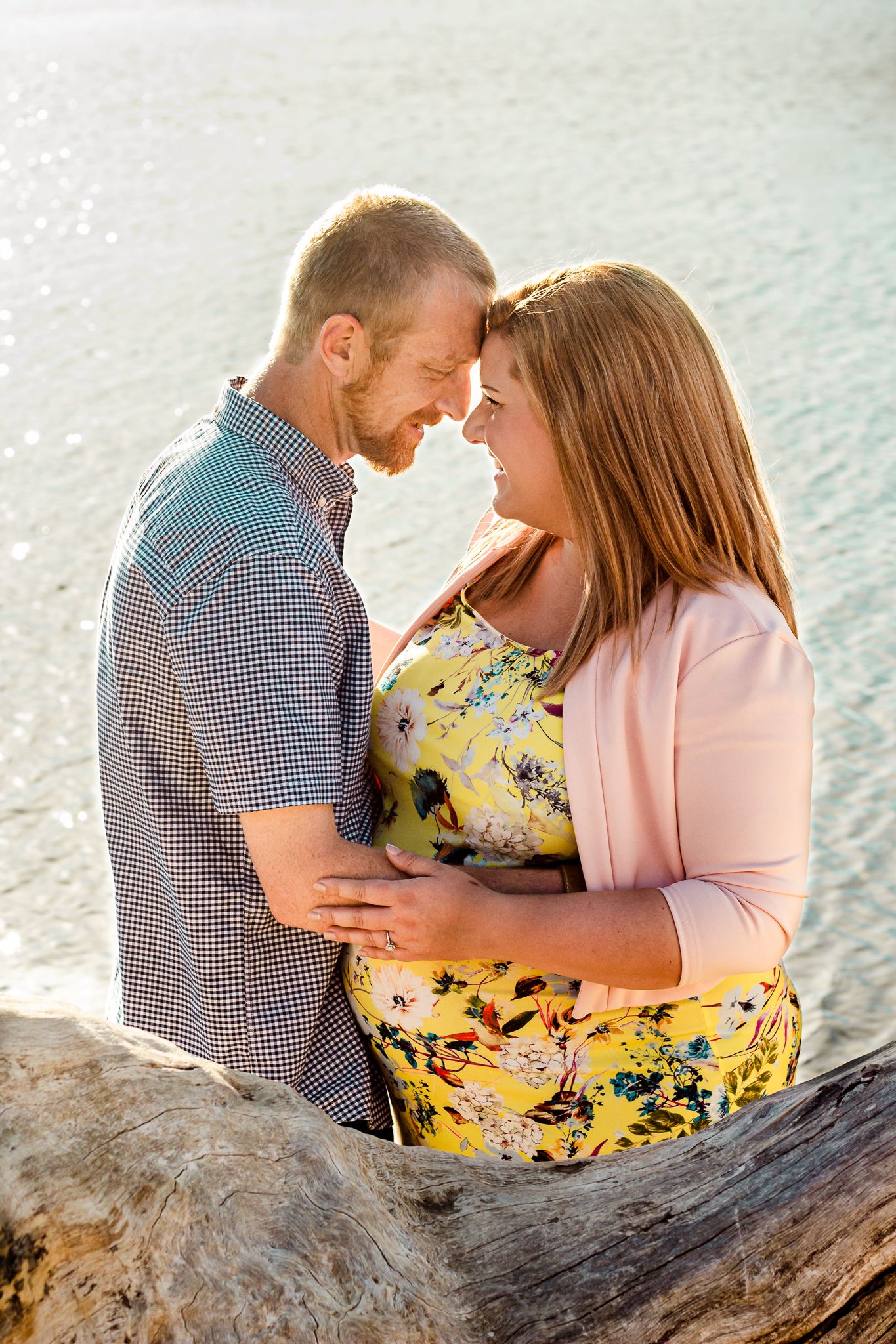 A maternity couple embrace and smile at each other by the sea on a Hayling Island Beach while the sun sets behind them.