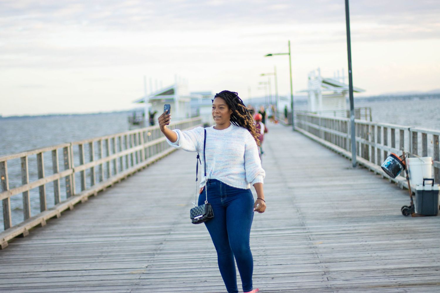 Person walking on wooden pier by ocean taking selfie while wearing casual white top and jeans.