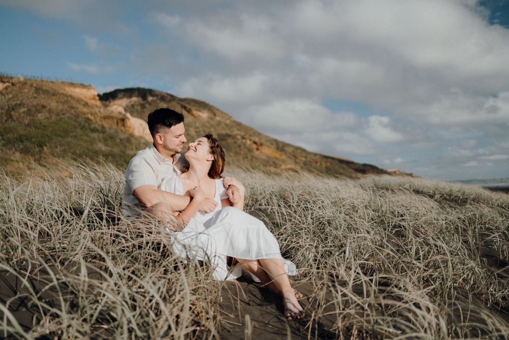 A romantic couple sits together in tall grass on a coastal hillside during sunset.