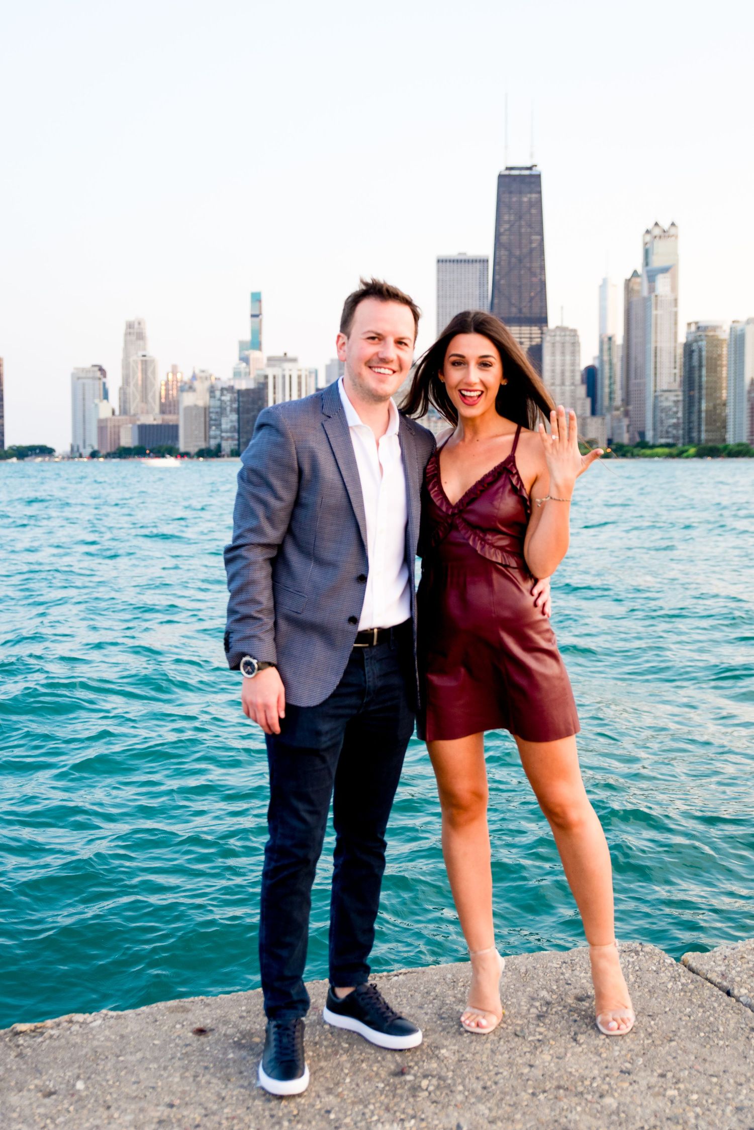 woman in maroon leather dress holding up her hand showing she is engaged in front of Chicago skyline at North Ave Beach