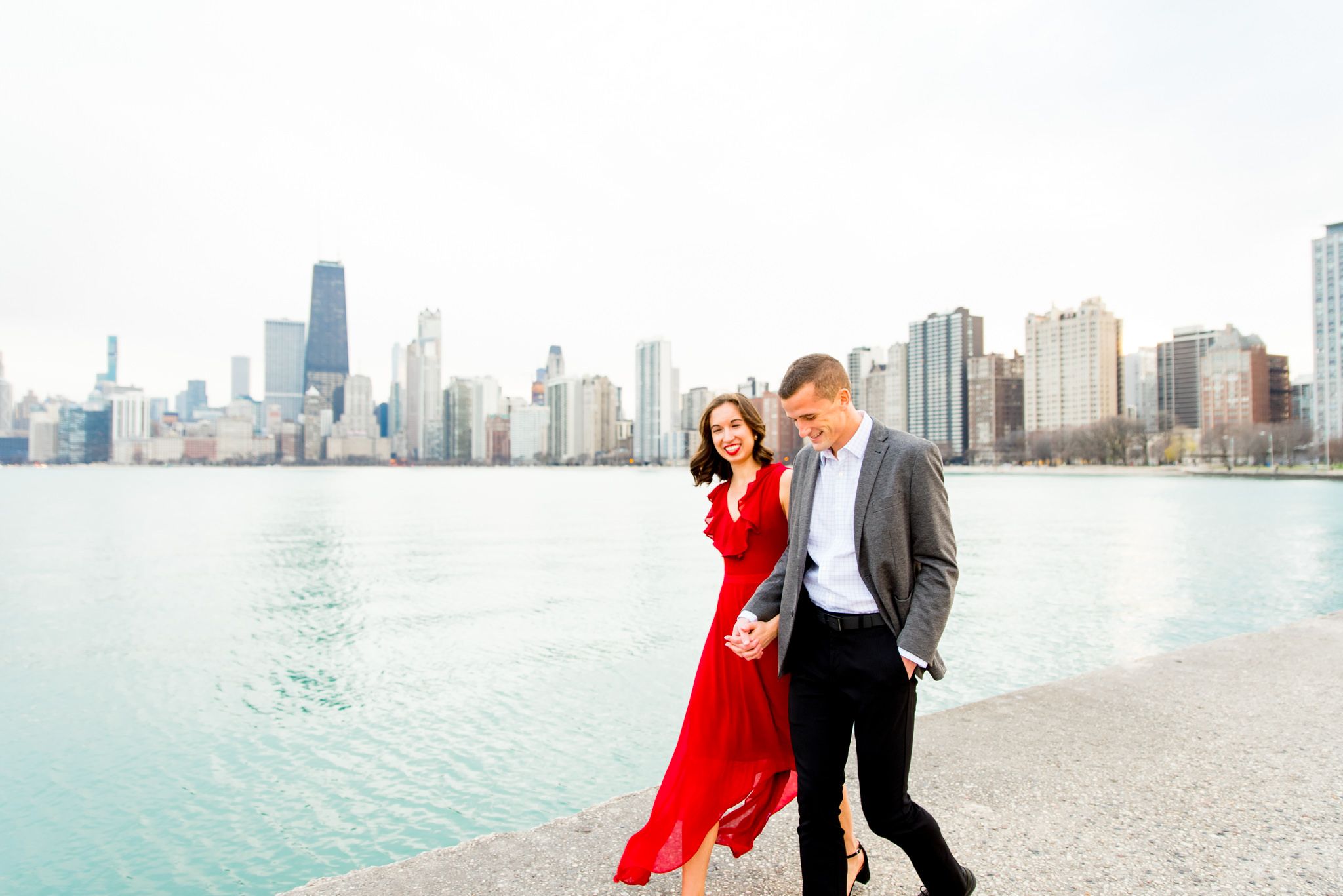 woman in red dress holding hands walking with man in grey suit along North Avenue Beach Chicago engagement pictures