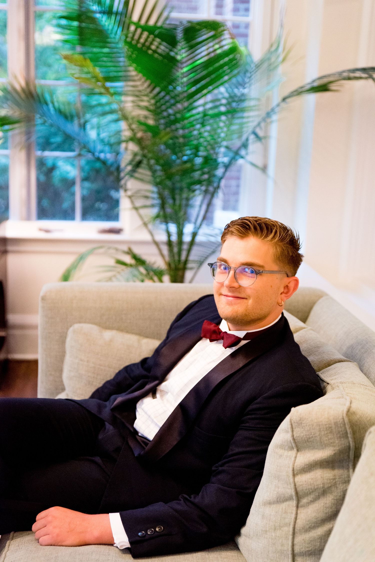blonde groom in navy suit and red bowtie and glasses smiling and sitting in the Woman's Club of Evanston sunroom
