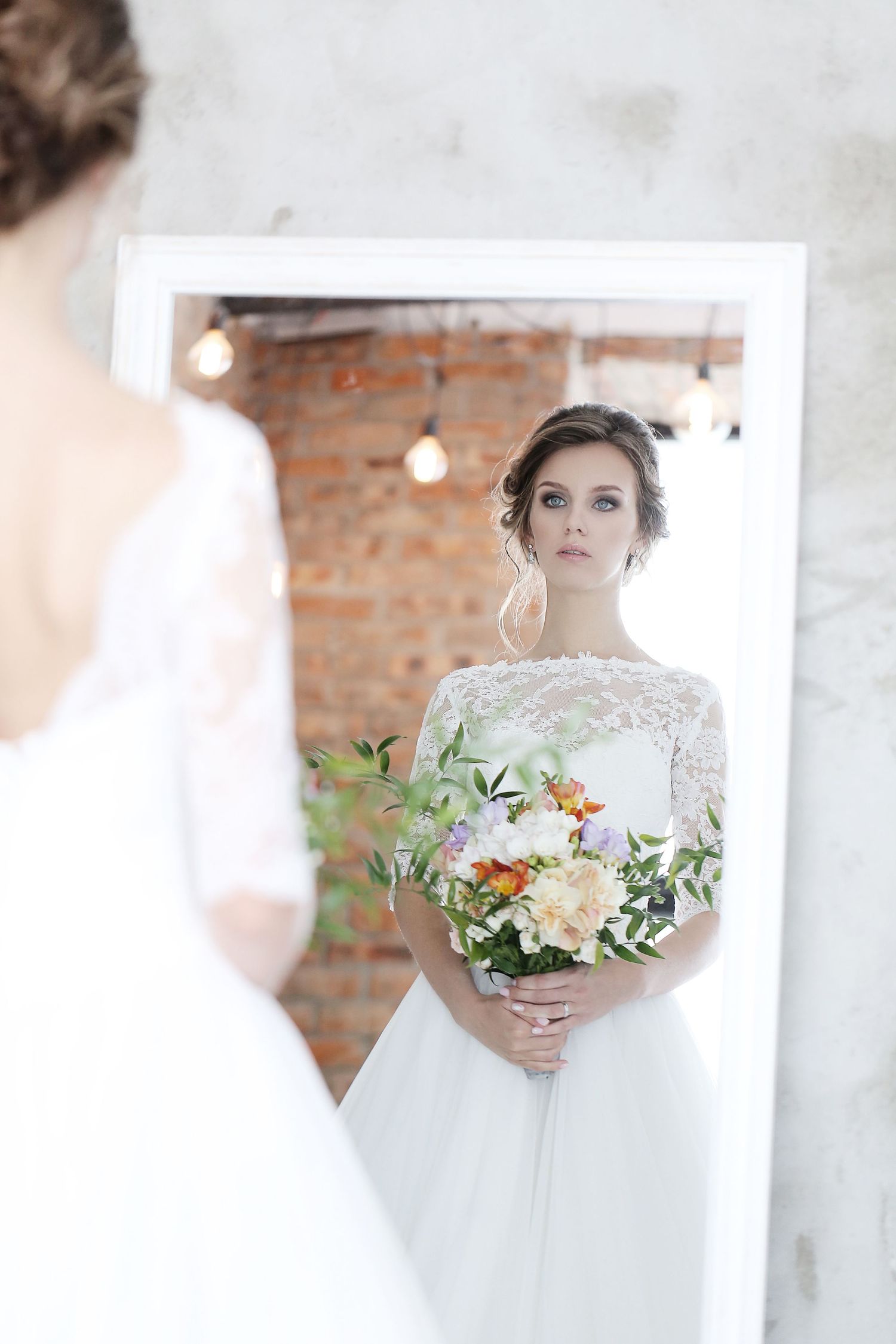Bride with floral bouquet gazes at reflection in mirror against exposed brick wall.
