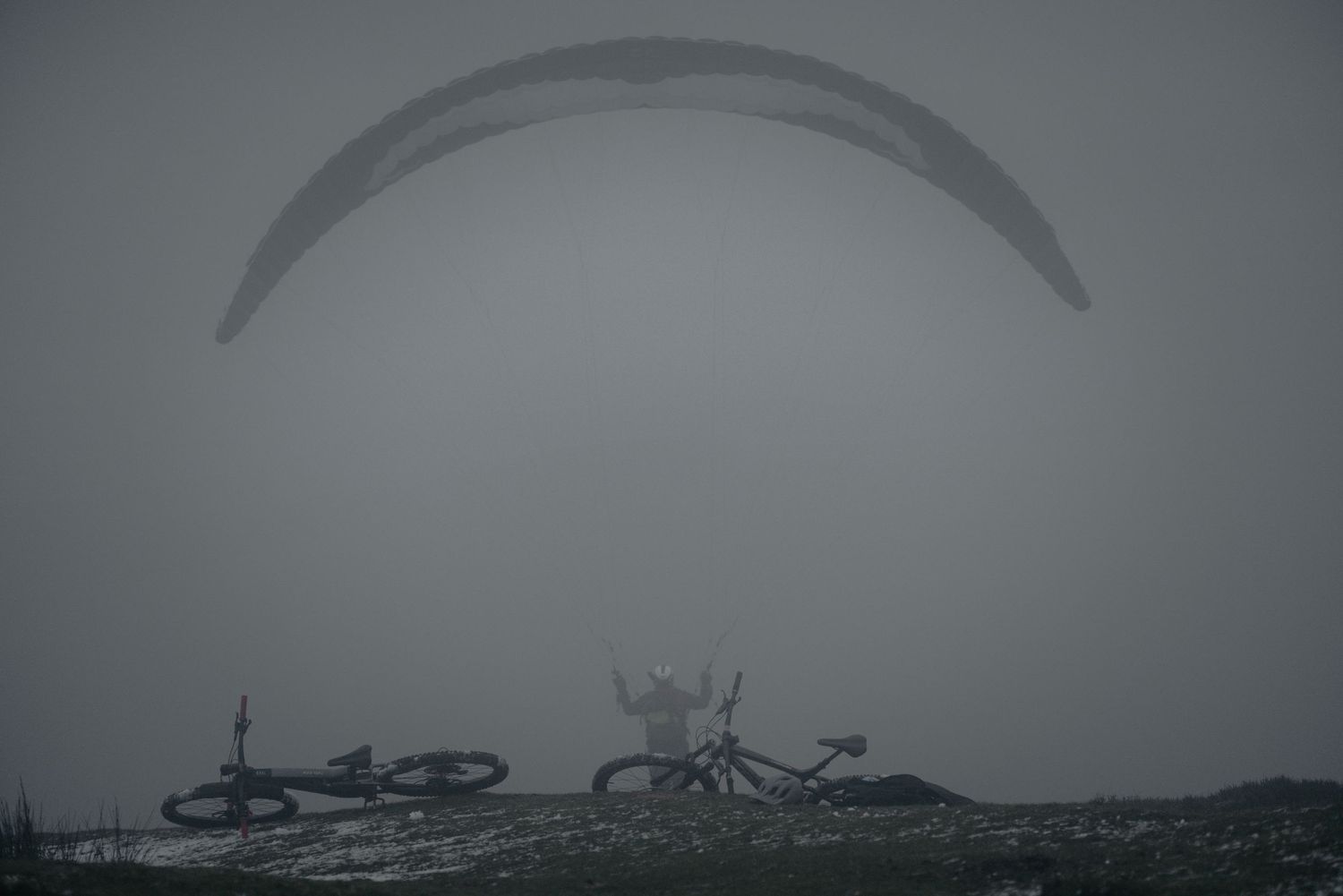 Silhouette of paraglider wing above parked bicycles in foggy conditions.