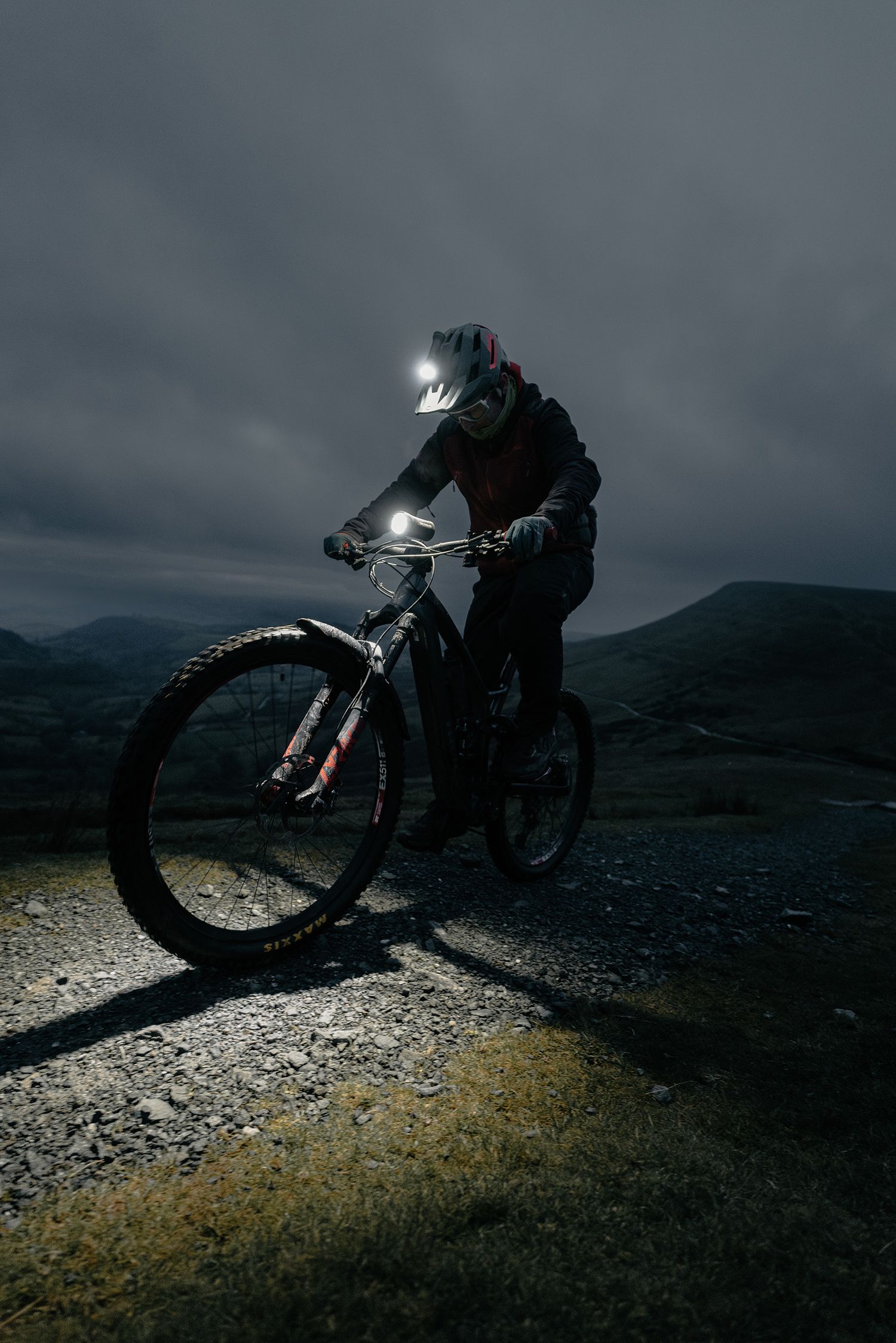 A mountain biker with headlamp rides on rocky terrain under dramatic stormy skies.