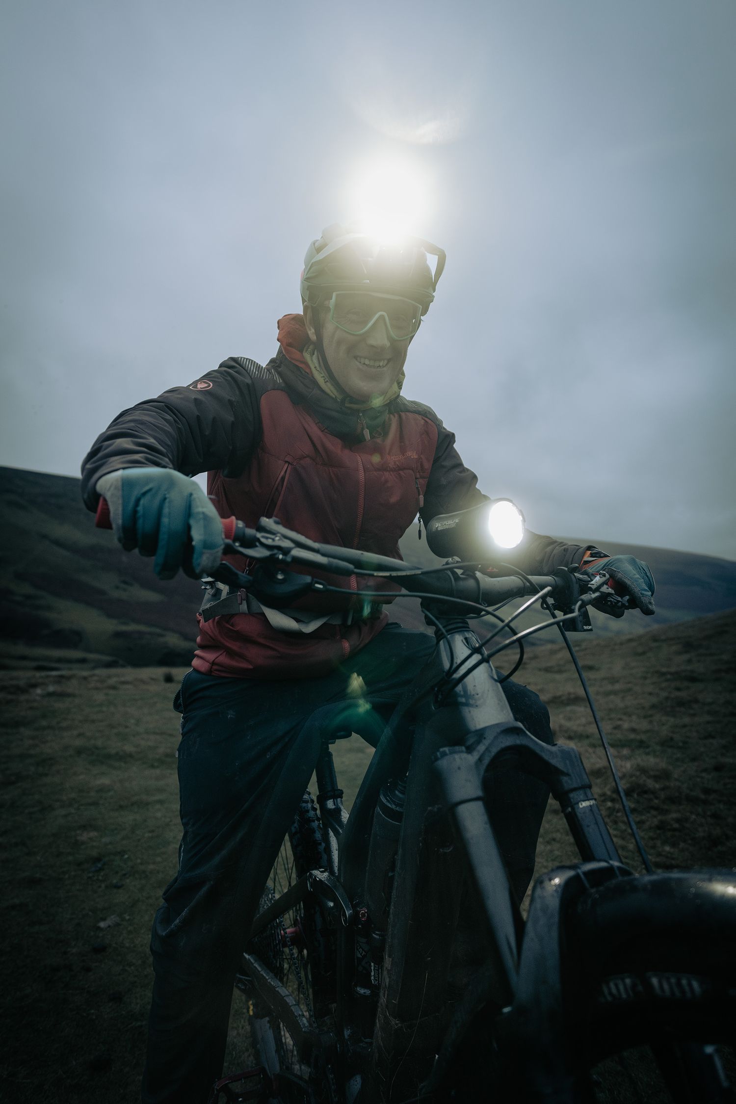 A mountain biker with headlamp rides in low light conditions on a hillside trail.