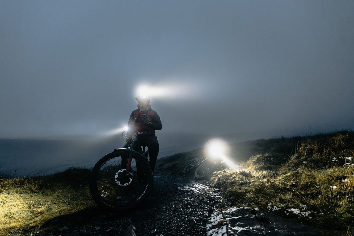 A mountain biker's headlamp creates dramatic lighting on a dark mountain trail at night.