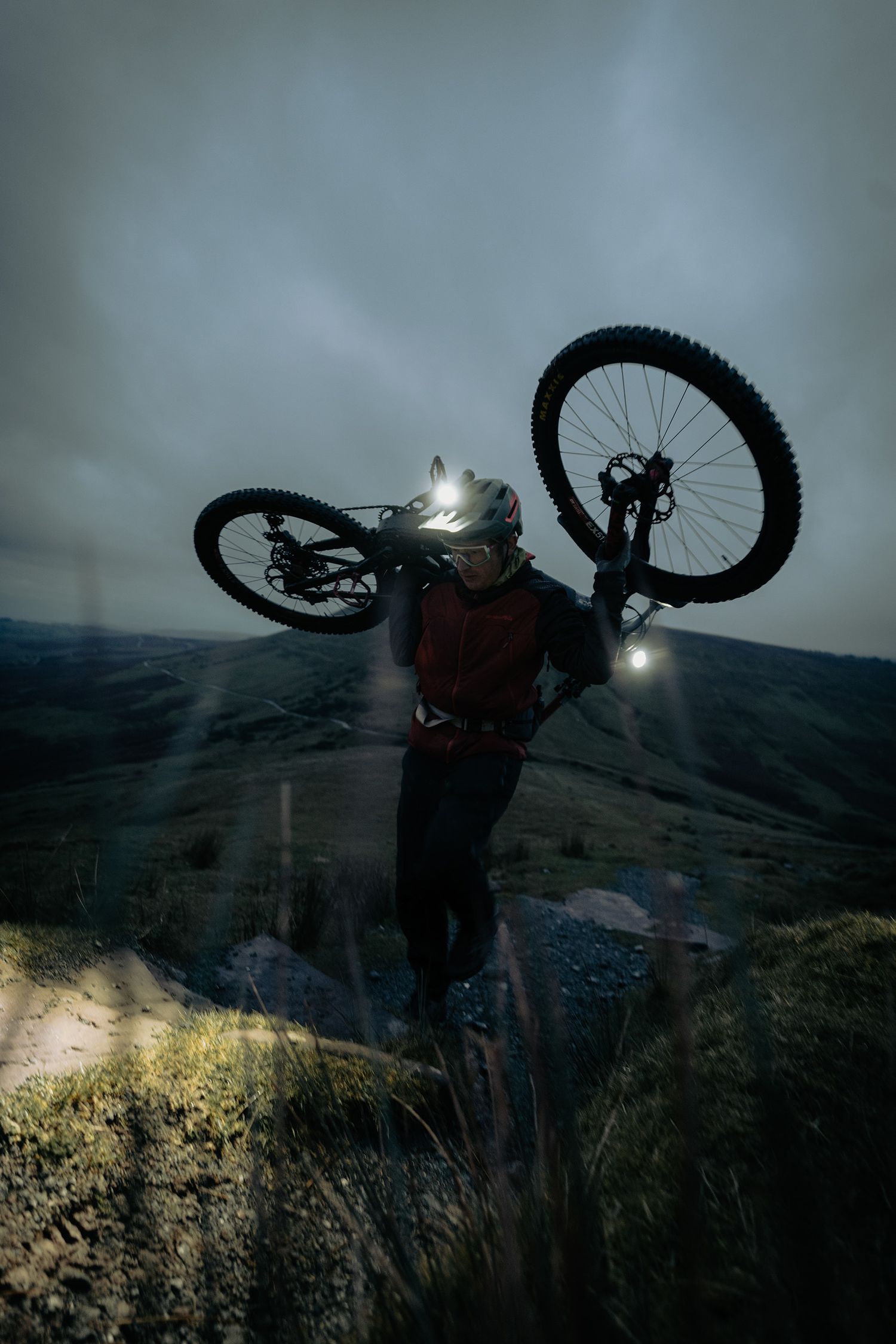 A mountain biker holds their bike overhead on a hillside at dusk with headlamps glowing.