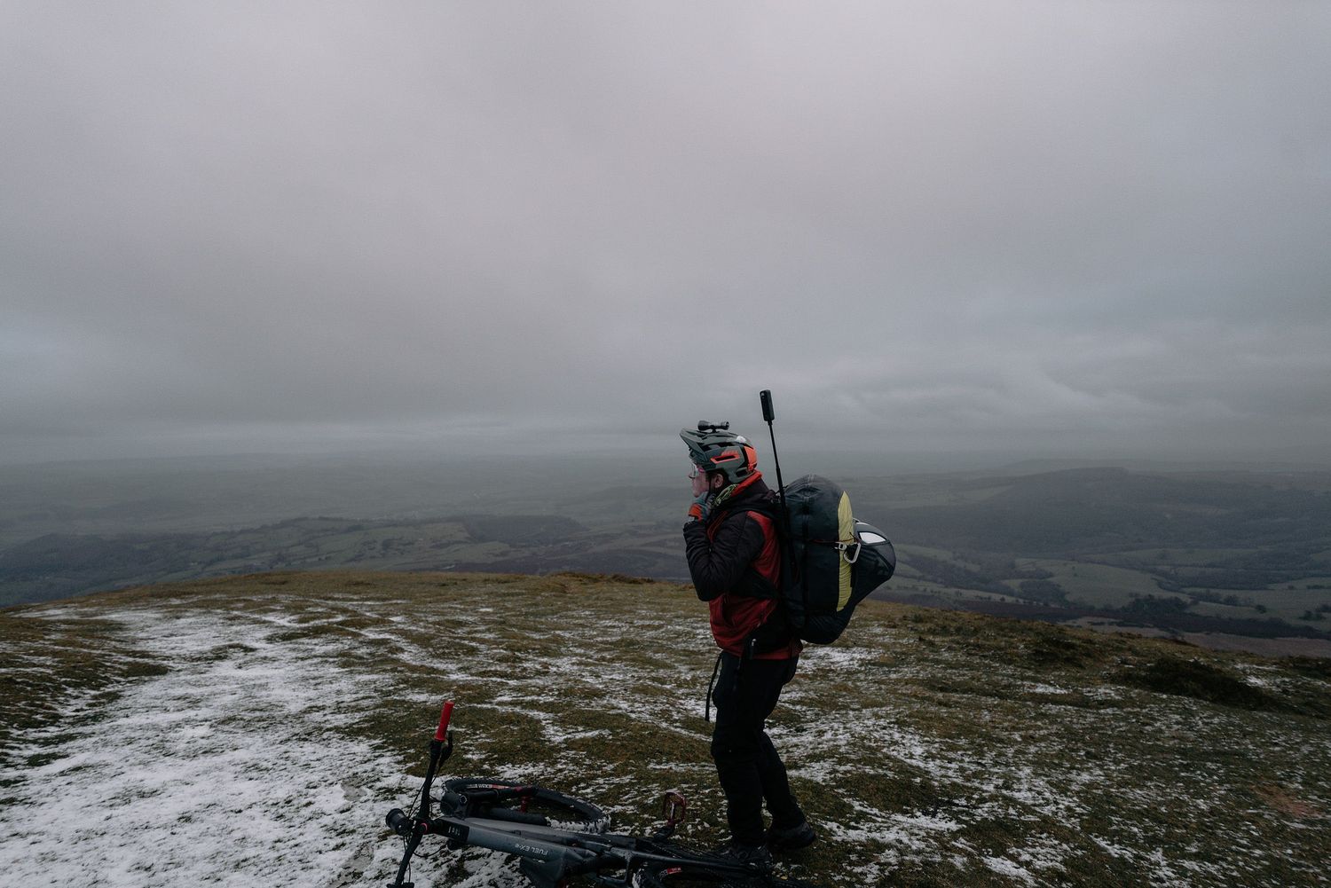 A mountain biker stands silhouetted against cloudy skies on a snowy hilltop with hiking gear.