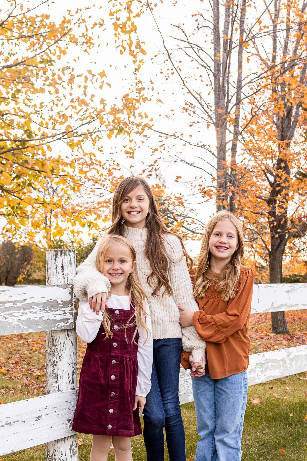 three sisters leaning against a old white fence with fall leaves falling in the background