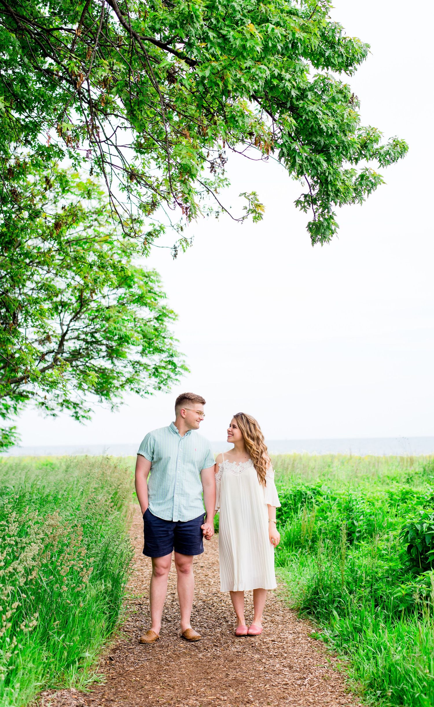 engaged couple in white dress and blue shirt hold hands and look at each other under a big green tree in the summer