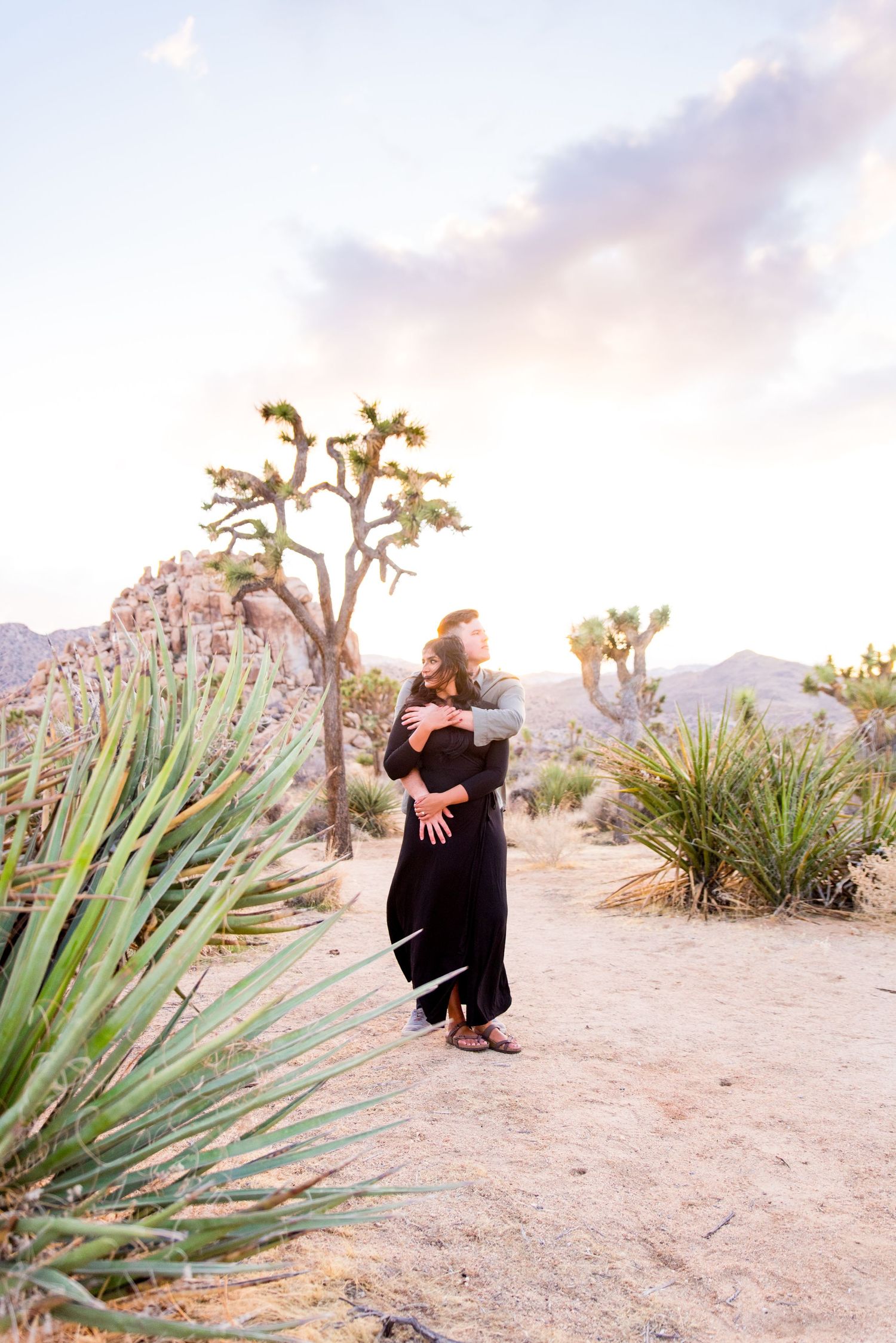 man in grey shirt hugs his fiancee in black dress from behind in Joshua Tree National Park for engagement pictures