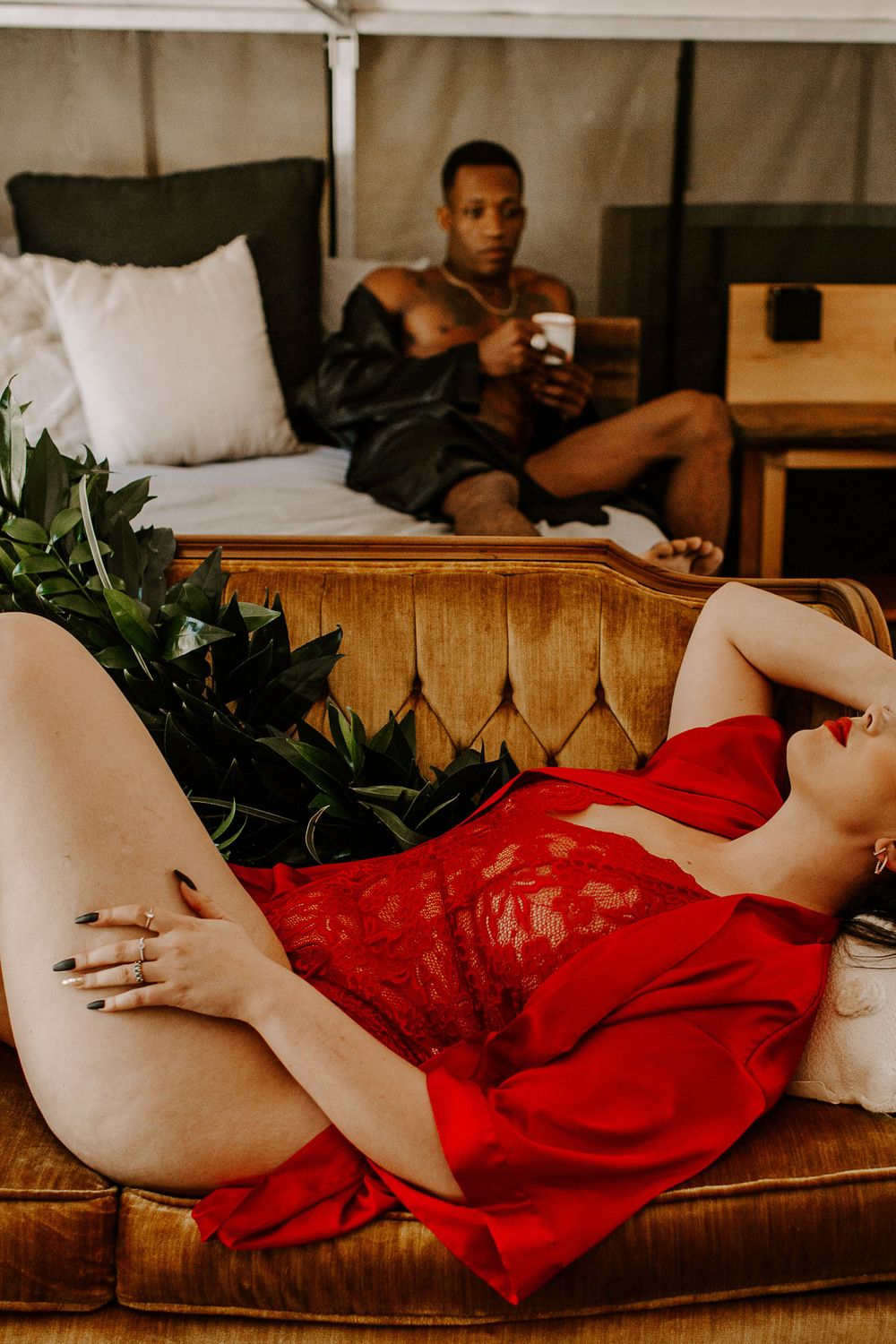 man sitting on a bed drinking coffee as woman lays on couch in lingerie during their couples session