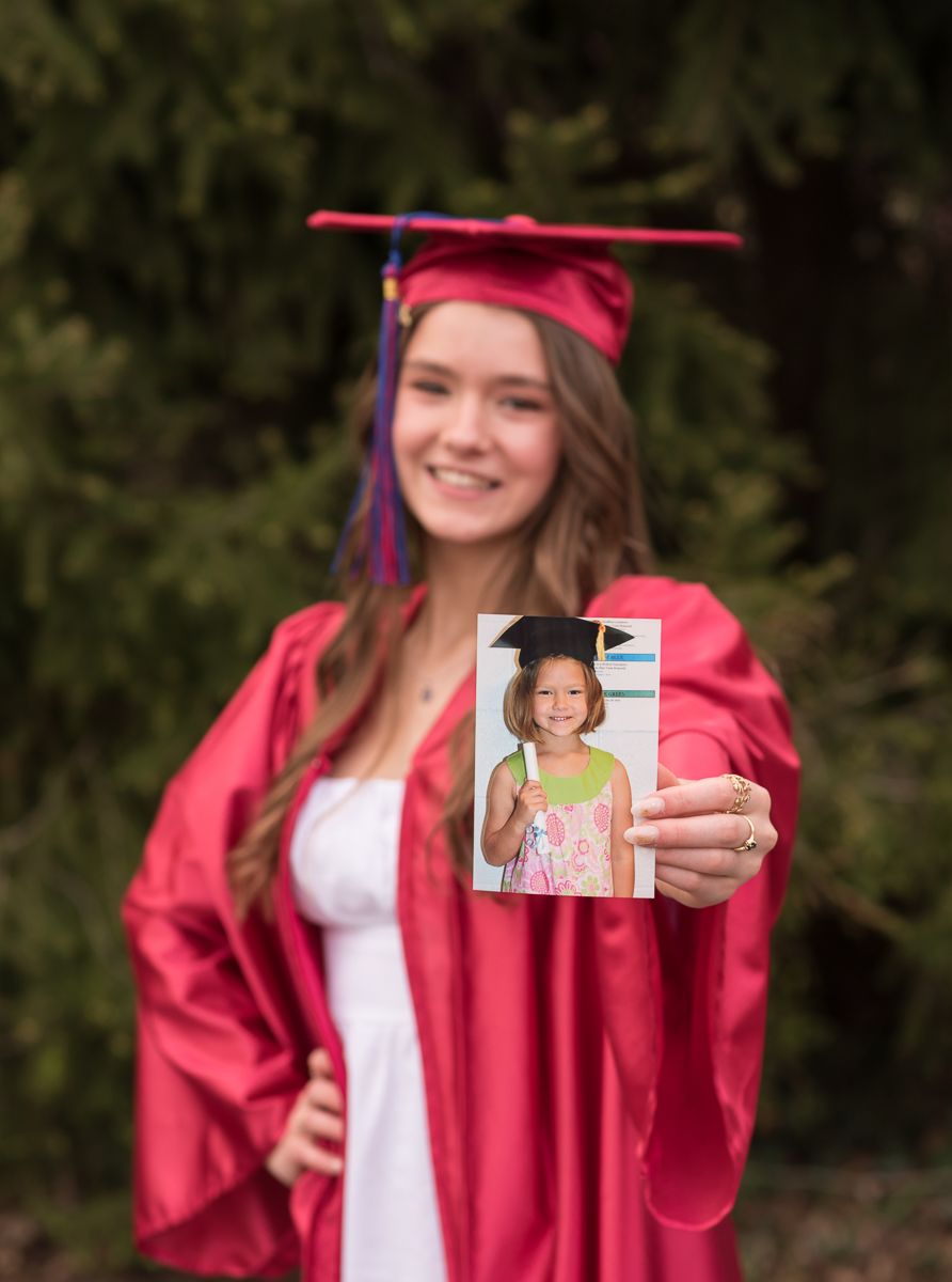 Portrait of an MHS grad in her red cap and gown, holding out a picture from kindergarten graduation, taken in Morgantown