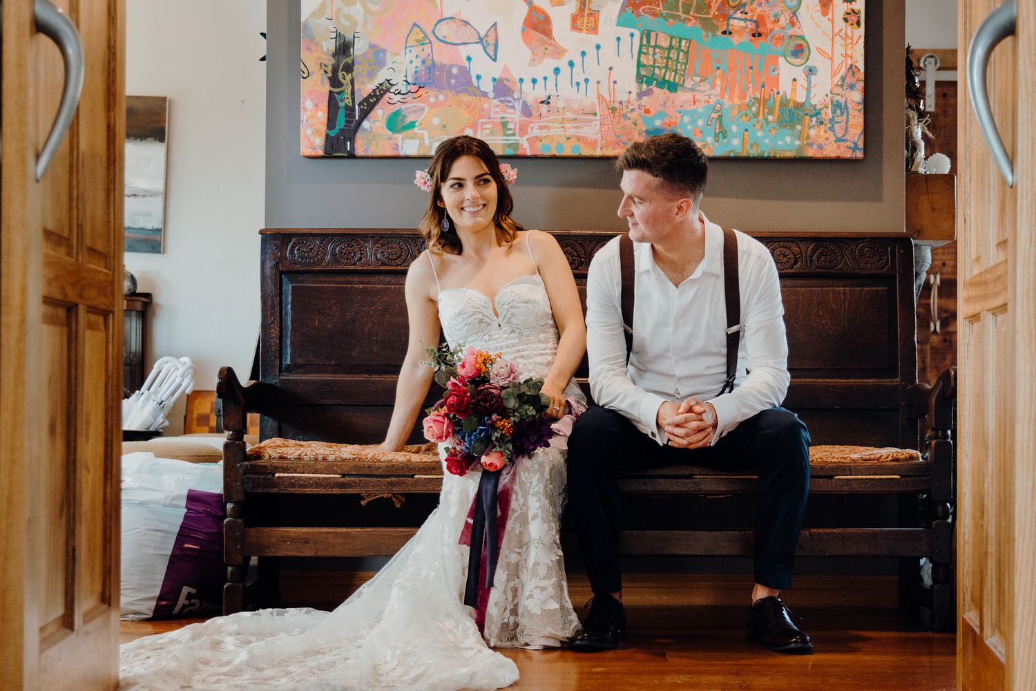 A couple sits on a wooden bench under colorful artwork in an intimate indoor setting.