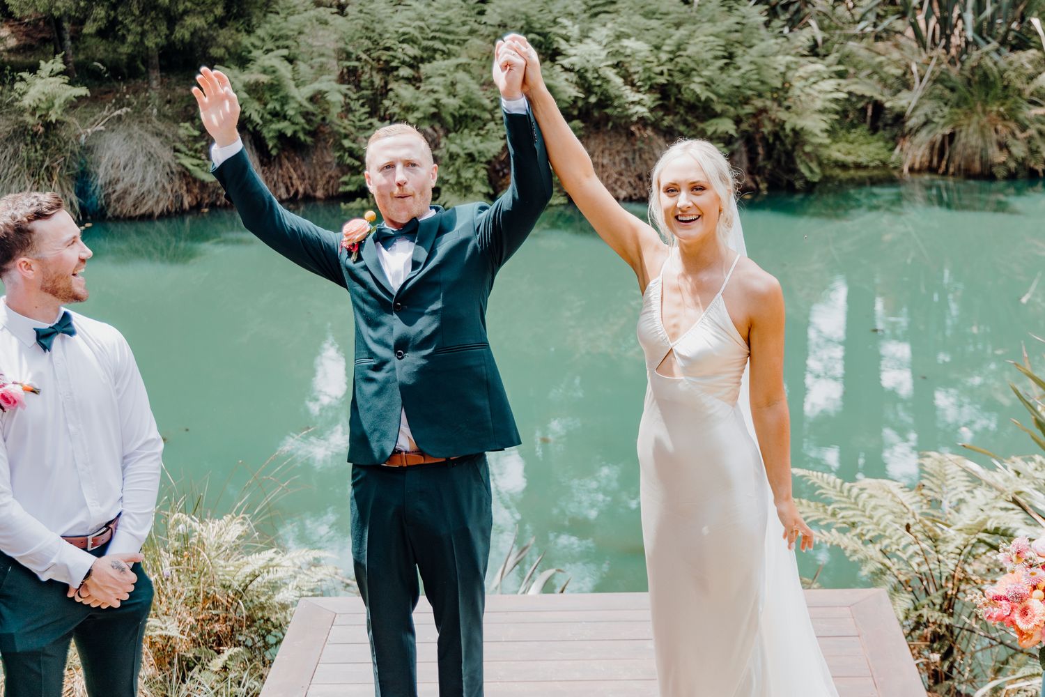 Newlyweds celebrate with raised arms on wooden deck overlooking turquoise waters.