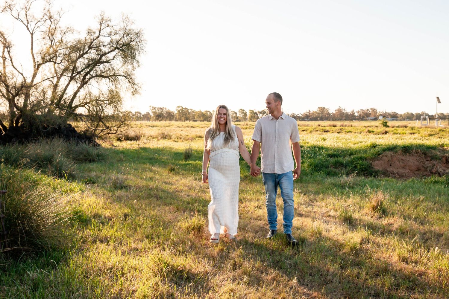 Couple stands together in a golden meadow during sunset with trees in background.
