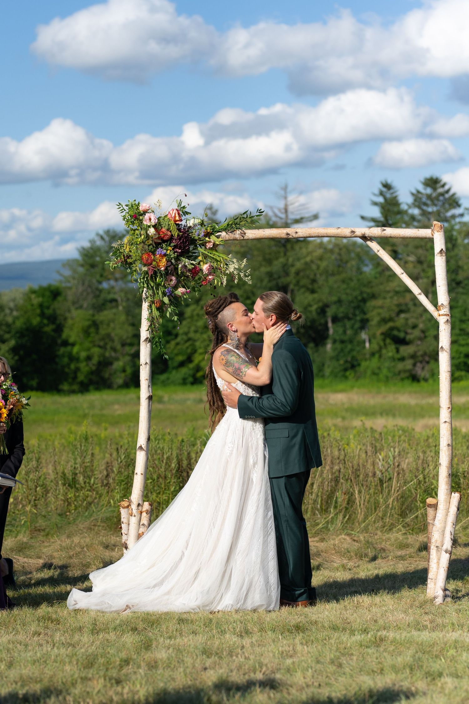 A couple shares their first kiss under a rustic wooden arch adorned with flowers in a scenic outdoor meadow.