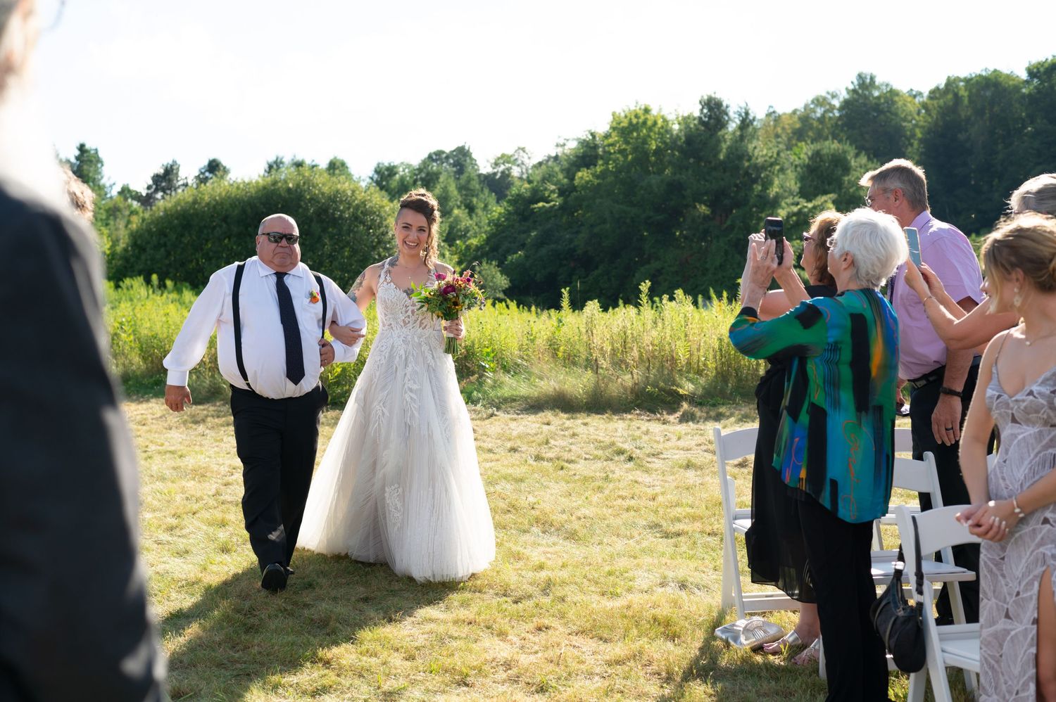 Outdoor wedding ceremony in sunny field with guests watching processional.