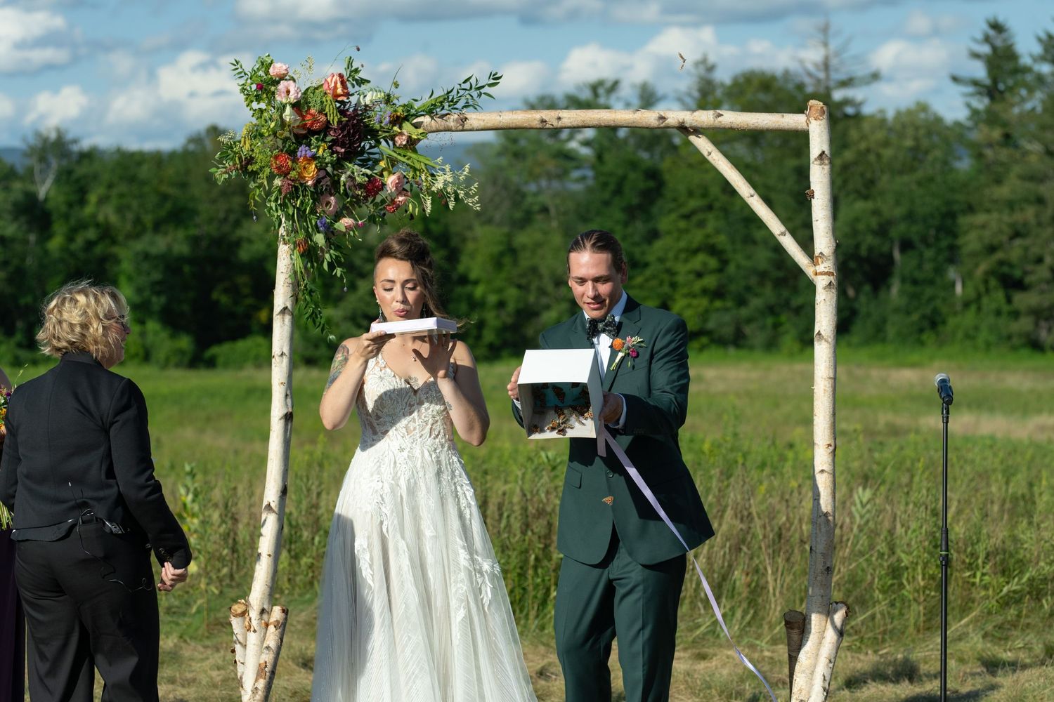 Outdoor wedding ceremony taking place under a birch wood arch decorated with colorful flowers.