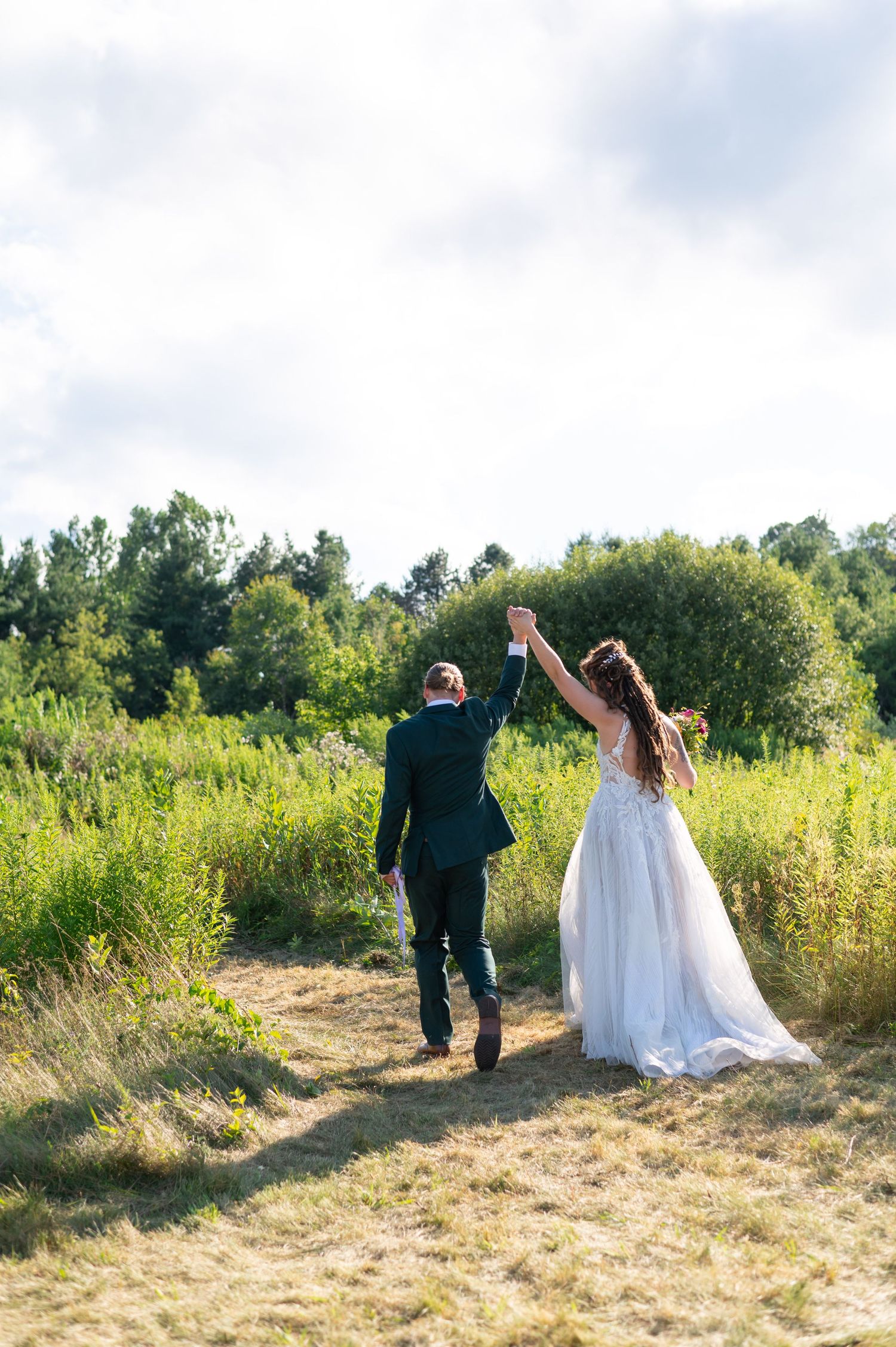 Newlyweds celebrate with raised hands while walking through a sunny meadow.