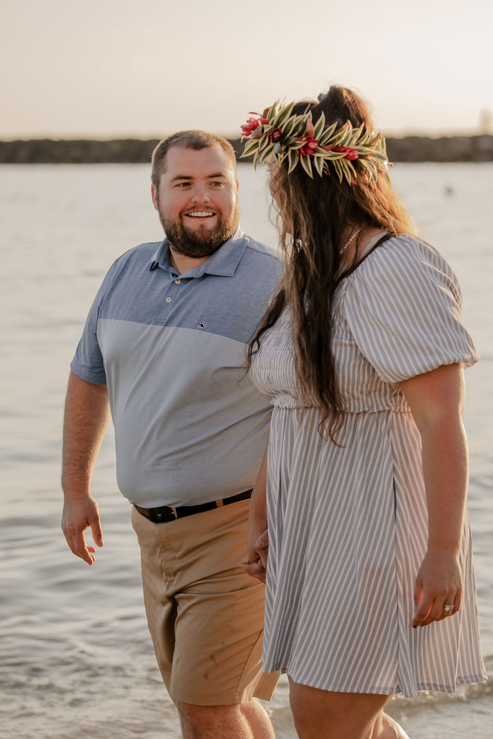 A couple shares a tender moment on the beach at sunset with one wearing a floral crown.