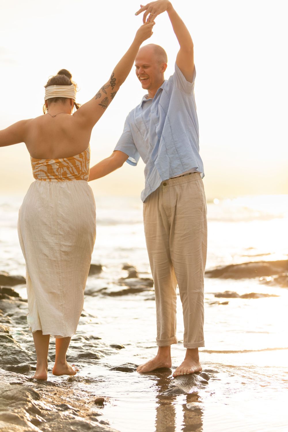 Couple dances playfully in ankle-deep water on rocky beach during golden hour.