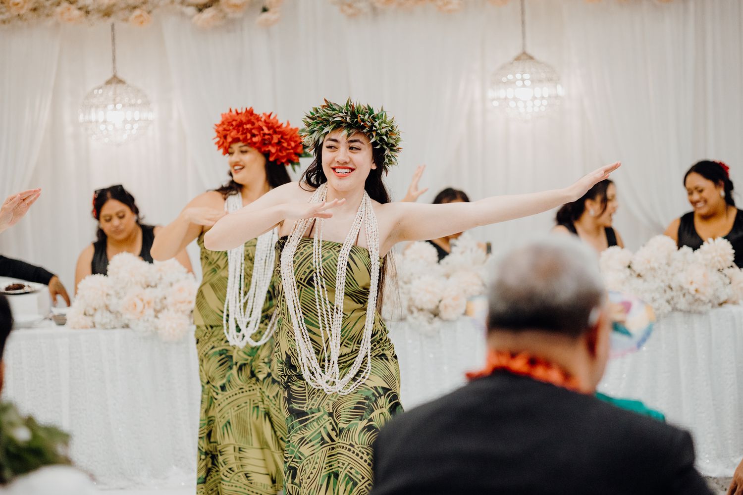 Performers in traditional Hawaiian attire dancing at a wedding reception with white backdrop.