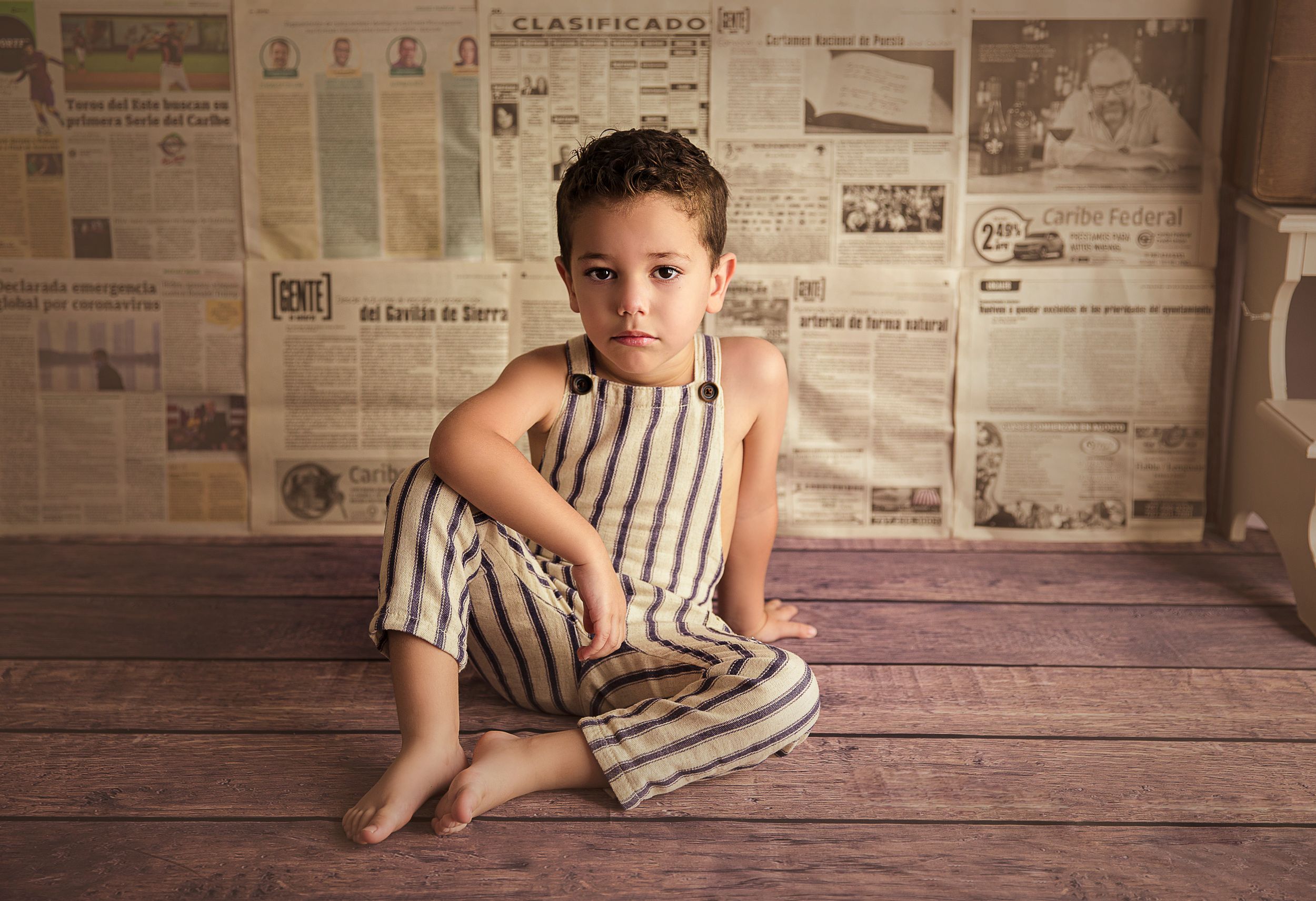 child sitting on wood floor wearing overalls newspaper backdrop
