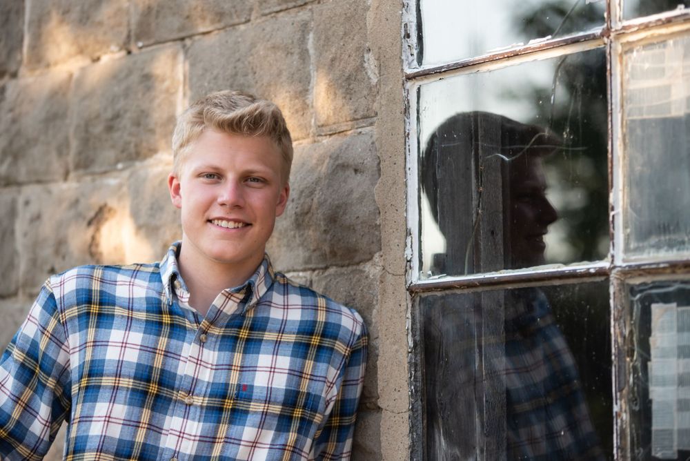 Blonde hair boy wearing blue plaid shirt standing in front of window of brick building