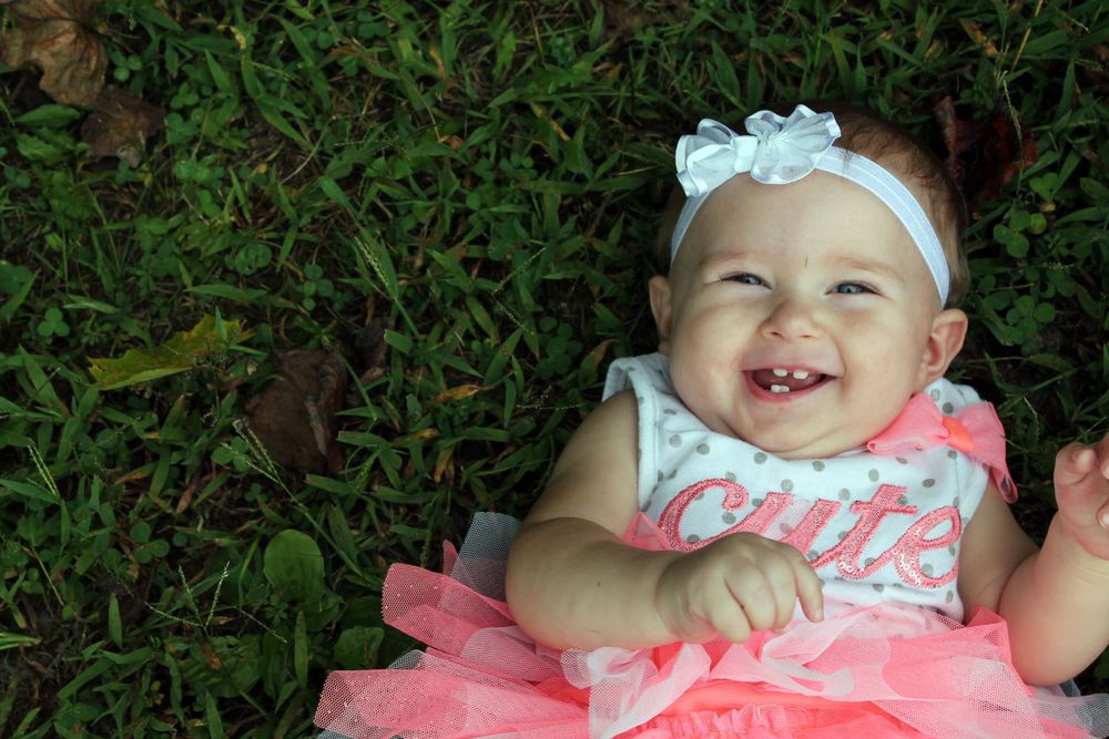 Baby wearing pink dress and white headband smiles while lying on green grass.