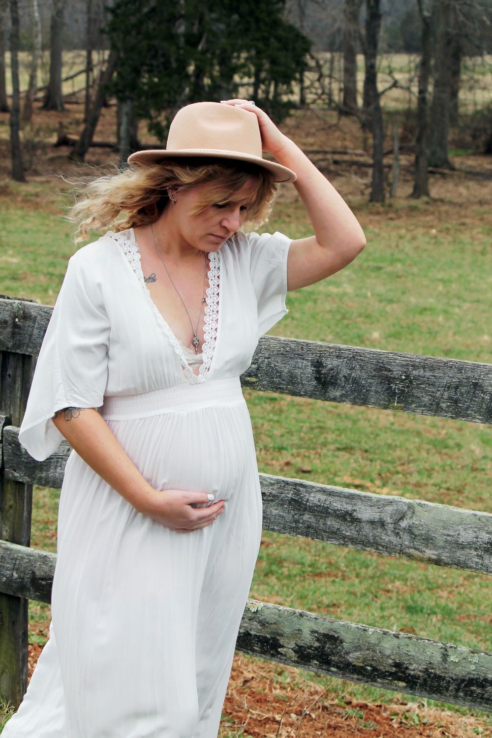 A woman in a white dress holding her hat poses by a rustic wooden fence.
