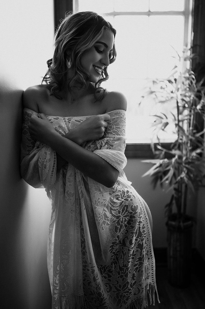 Artistic black and white photo of person in lace dress by window with soft natural lighting.