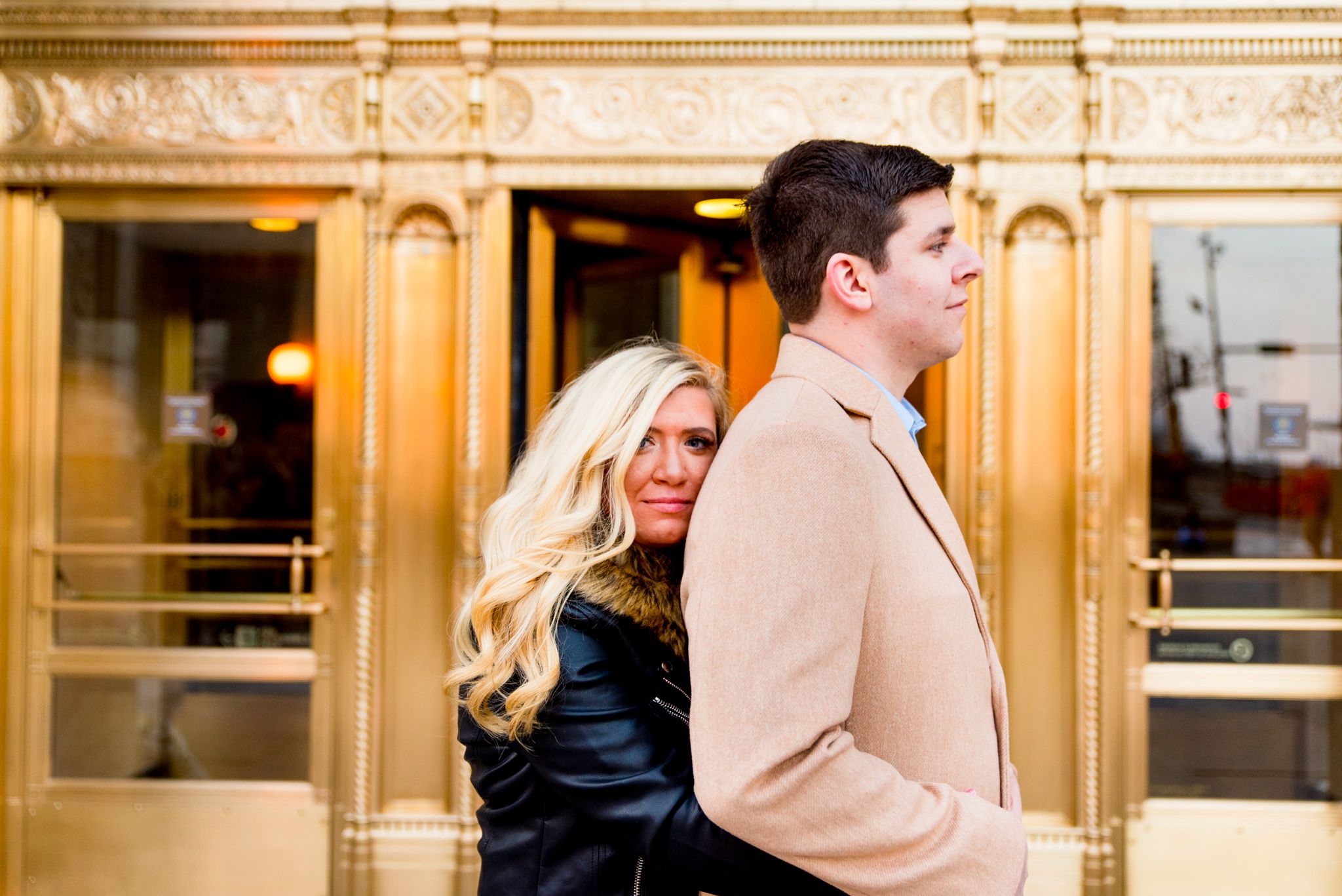 blonde woman in black coat hugging fiance from behind in front of gold doors at Wrigley Building Engagement pictures