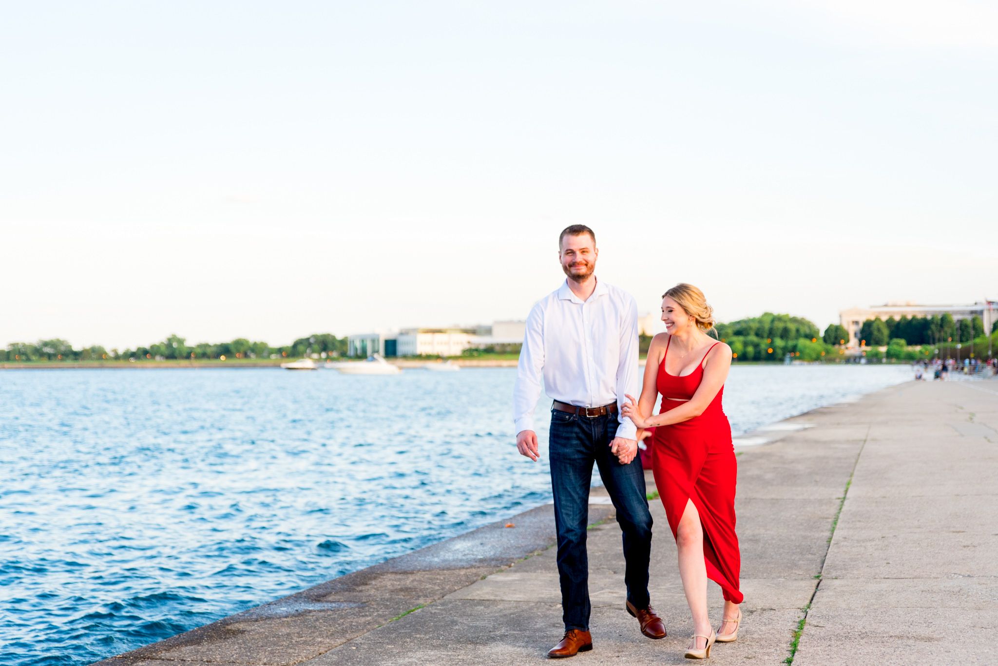 man in white shirt and woman in red dress walking along Chicago lakefront trail at sunset for engagement pictures