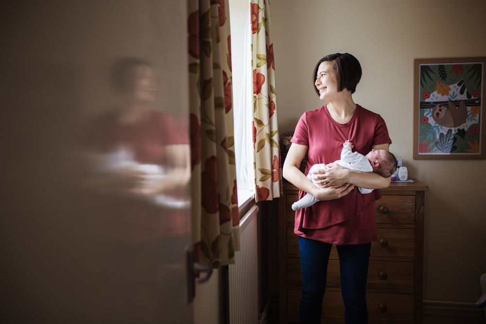 A mother holding her newborn baby looking out of a window at a newborn photography session by Hannah Veale Photography