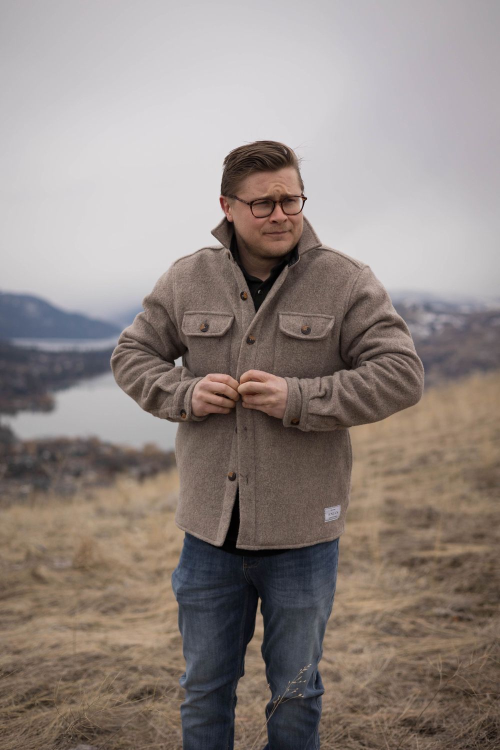 Person in tan fleece jacket and glasses standing on hillside with mountains in background.
