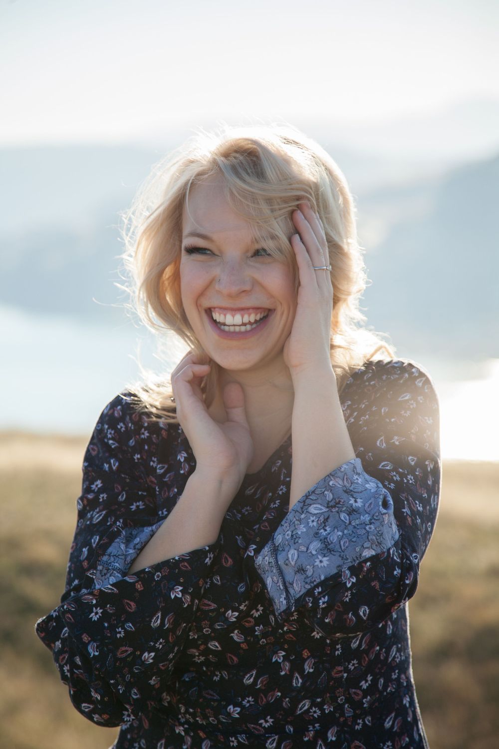 Portrait of woman in floral dress laughing with hands on face against mountain backdrop.