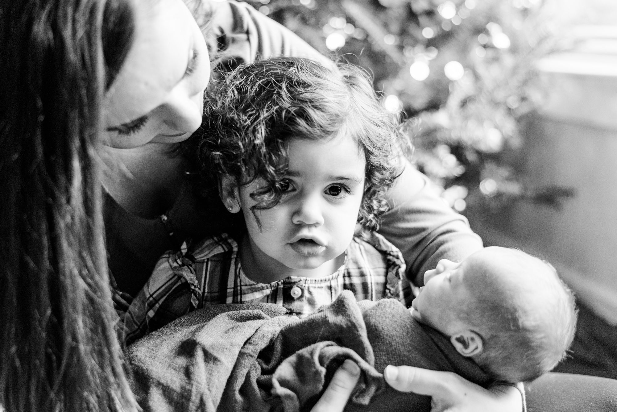 Lifestyle mother and big sister holding newborn baby in pink wrap in front of Christmas tree in Pittsburgh, Pennsylvania