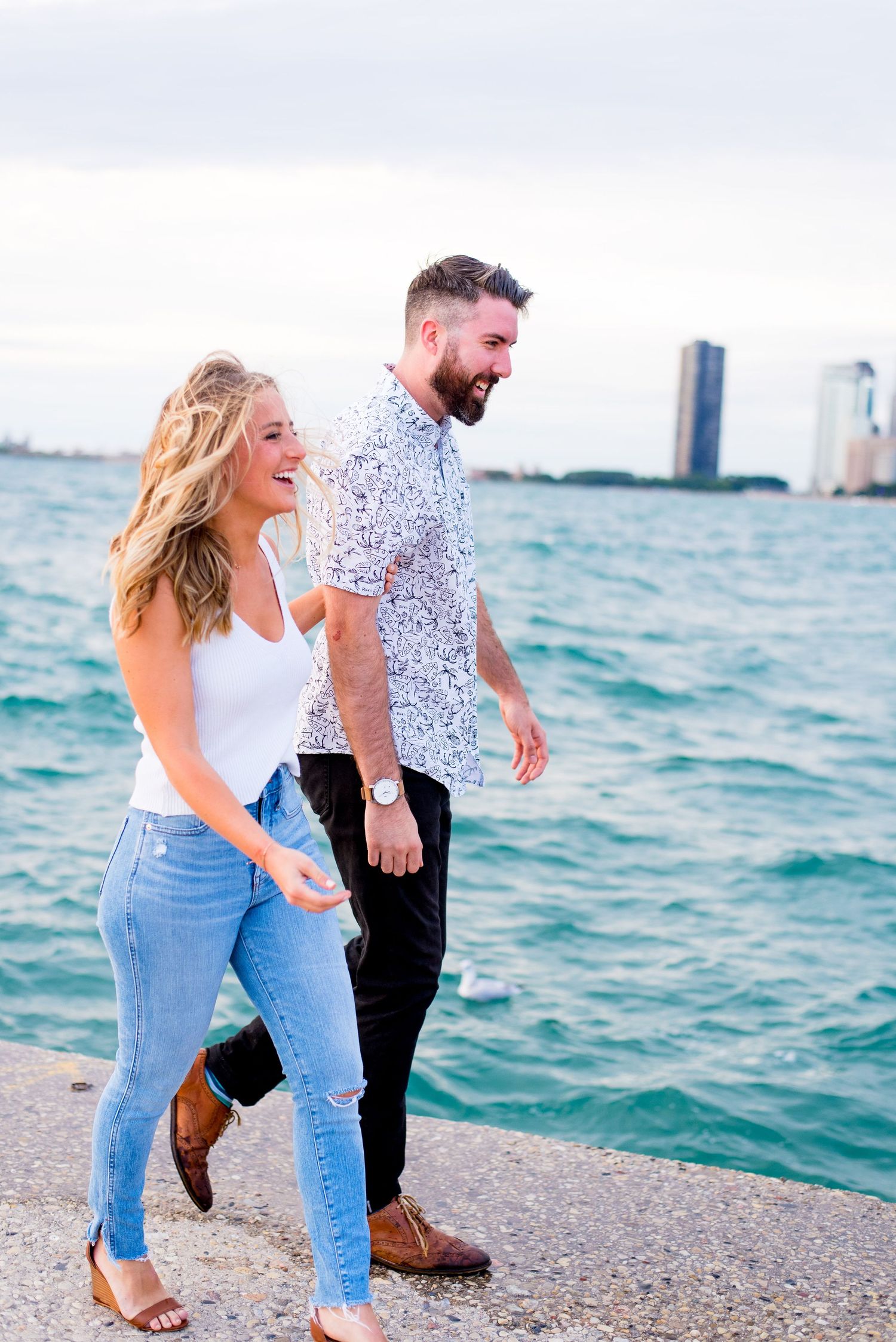 engaged man and woman link arms and laugh as they walk along the pier at North Avenue Beach in Chicago