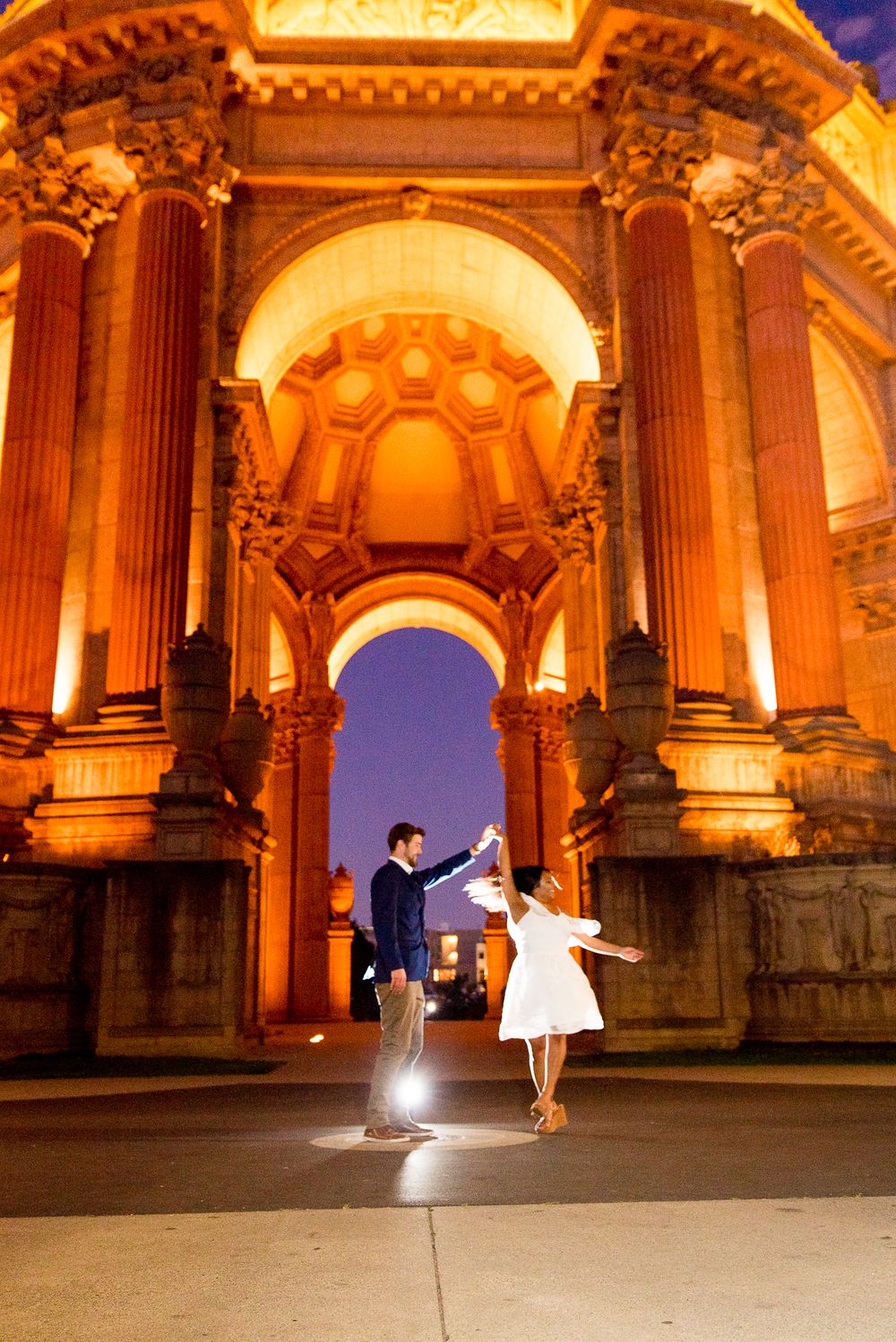 young couple in front of Palace of Fine Arts at night, lit up with blue sky in background