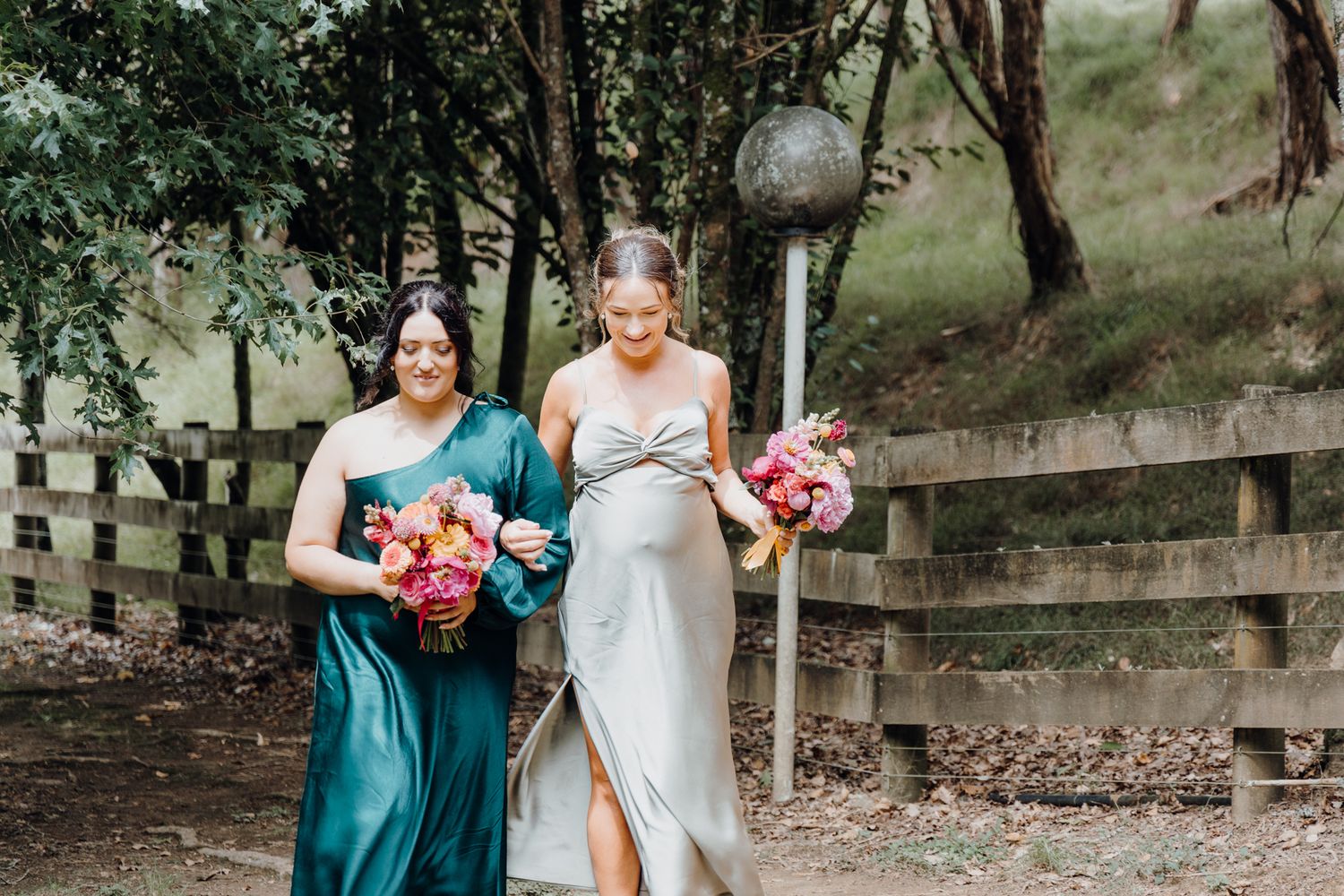 Attendants in teal and silver dresses carry vibrant floral bouquets near a wooden fence.