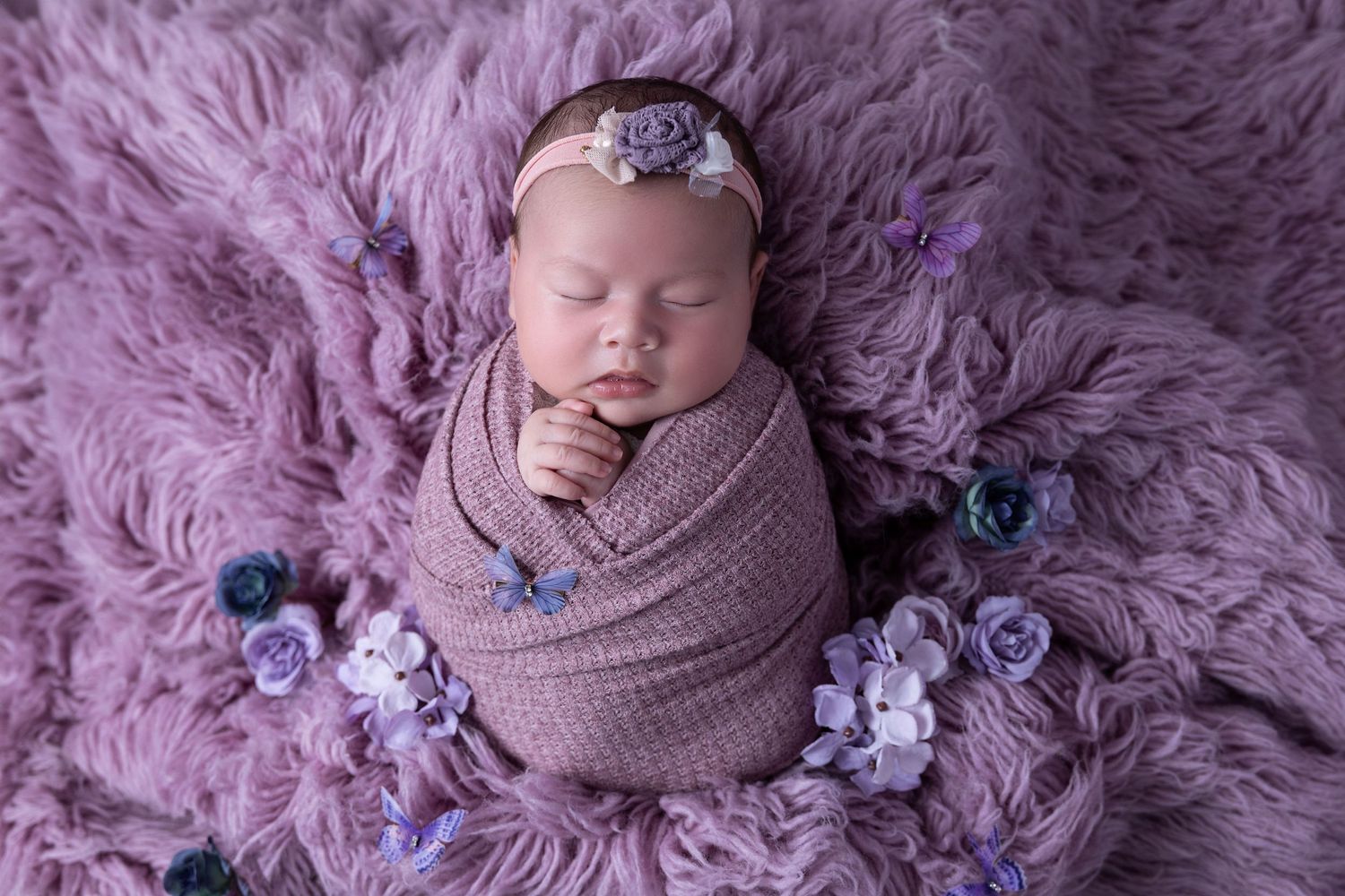 Newborn wrapped in pink swaddling cloth sleeps peacefully on purple fur blanket surrounded by purple flowers.