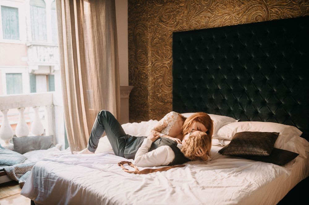 Couple resting on white bedding in moody indoor boudoir setting.