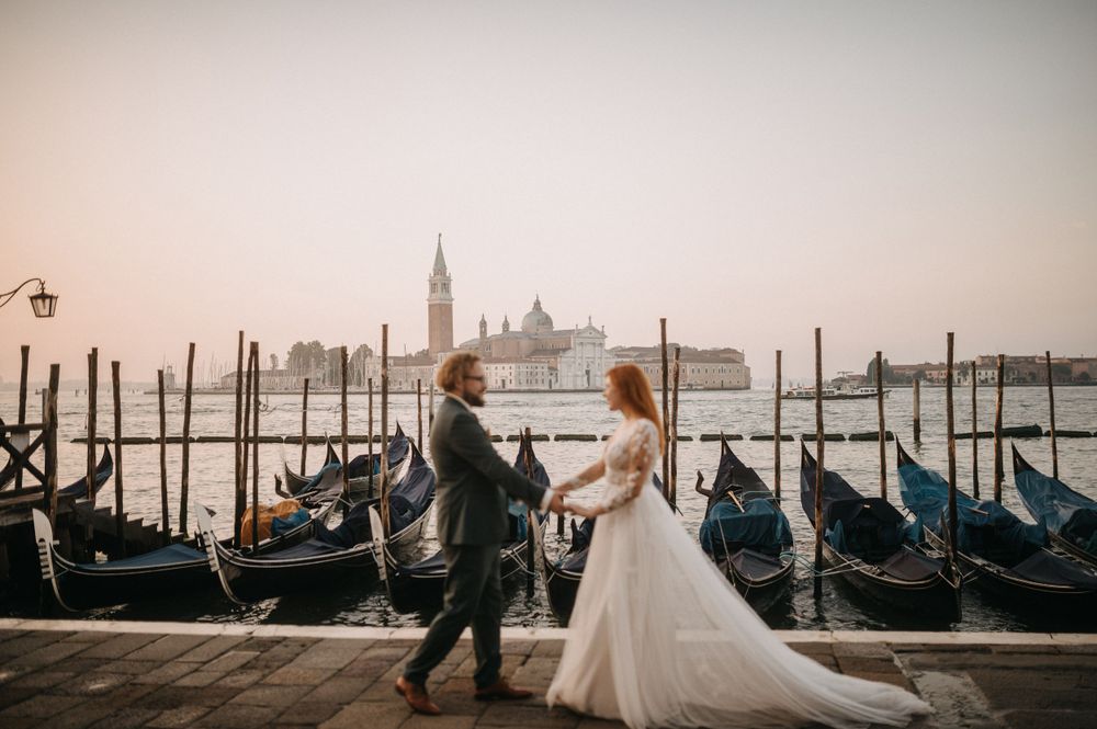 Couple in wedding attire dancing by gondolas with Venice skyline at dusk.