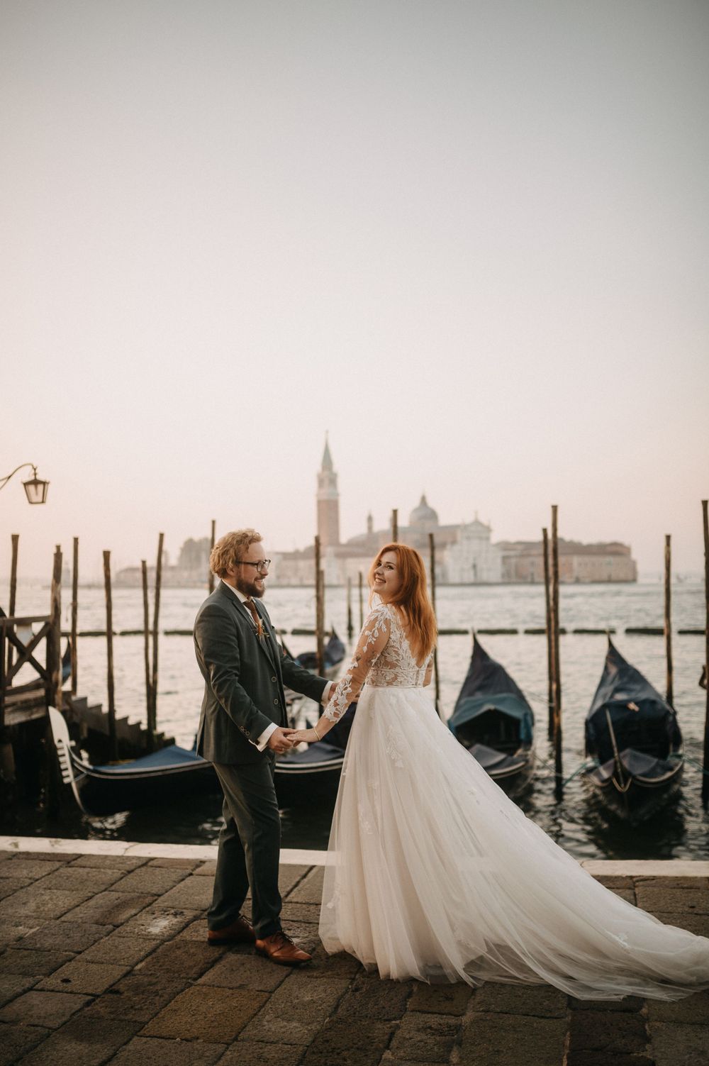 Wedding couple stands on Venice dock at sunset with iconic gondolas and cityscape in the background.