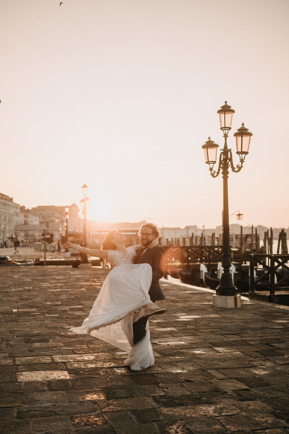 Dancing couple in wedding attire spin joyfully on Venice's cobblestone waterfront at sunset.
