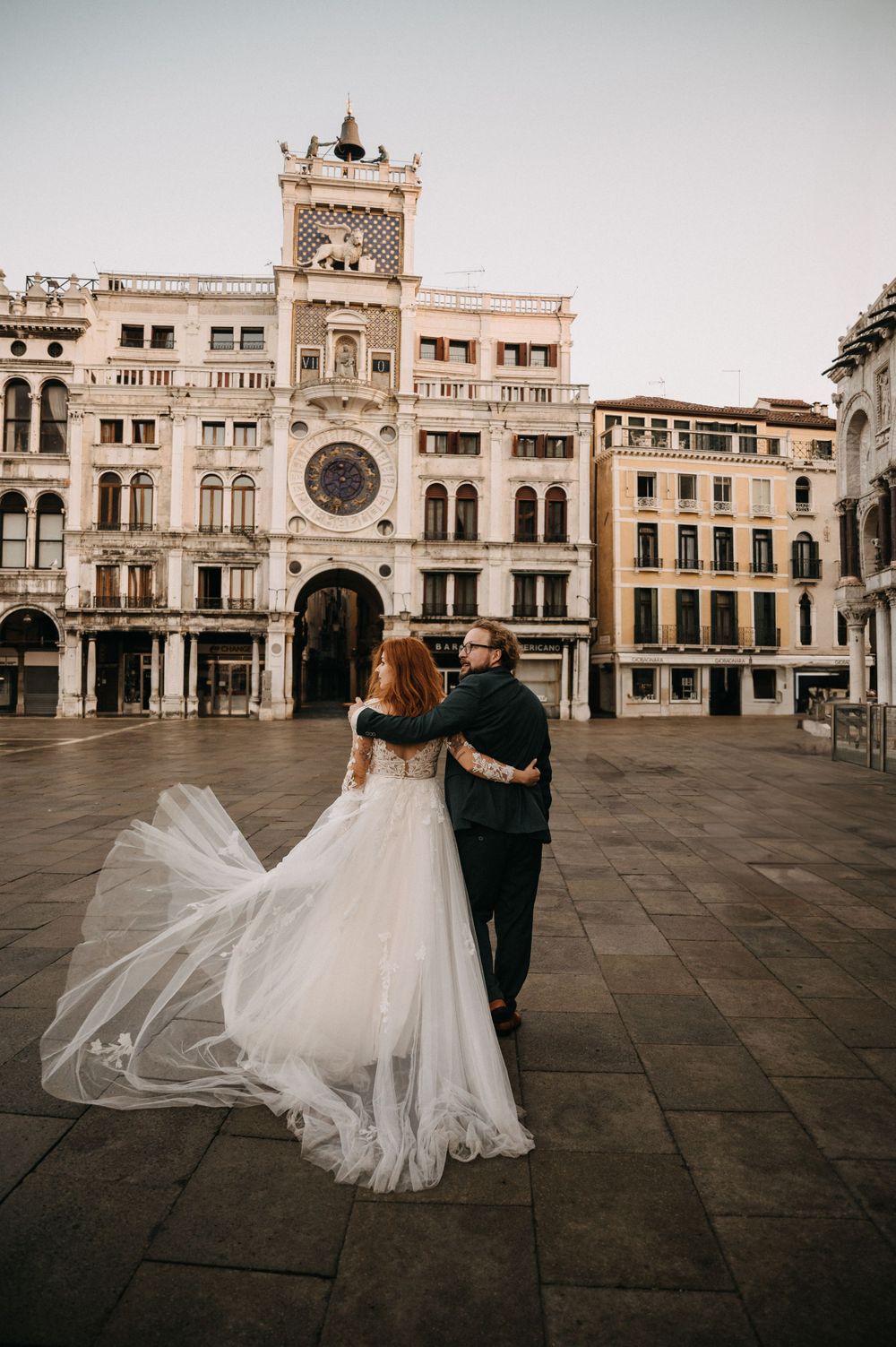Elegant bride and groom walk across Venice's St. Mark's Square with historic architecture behind them.
