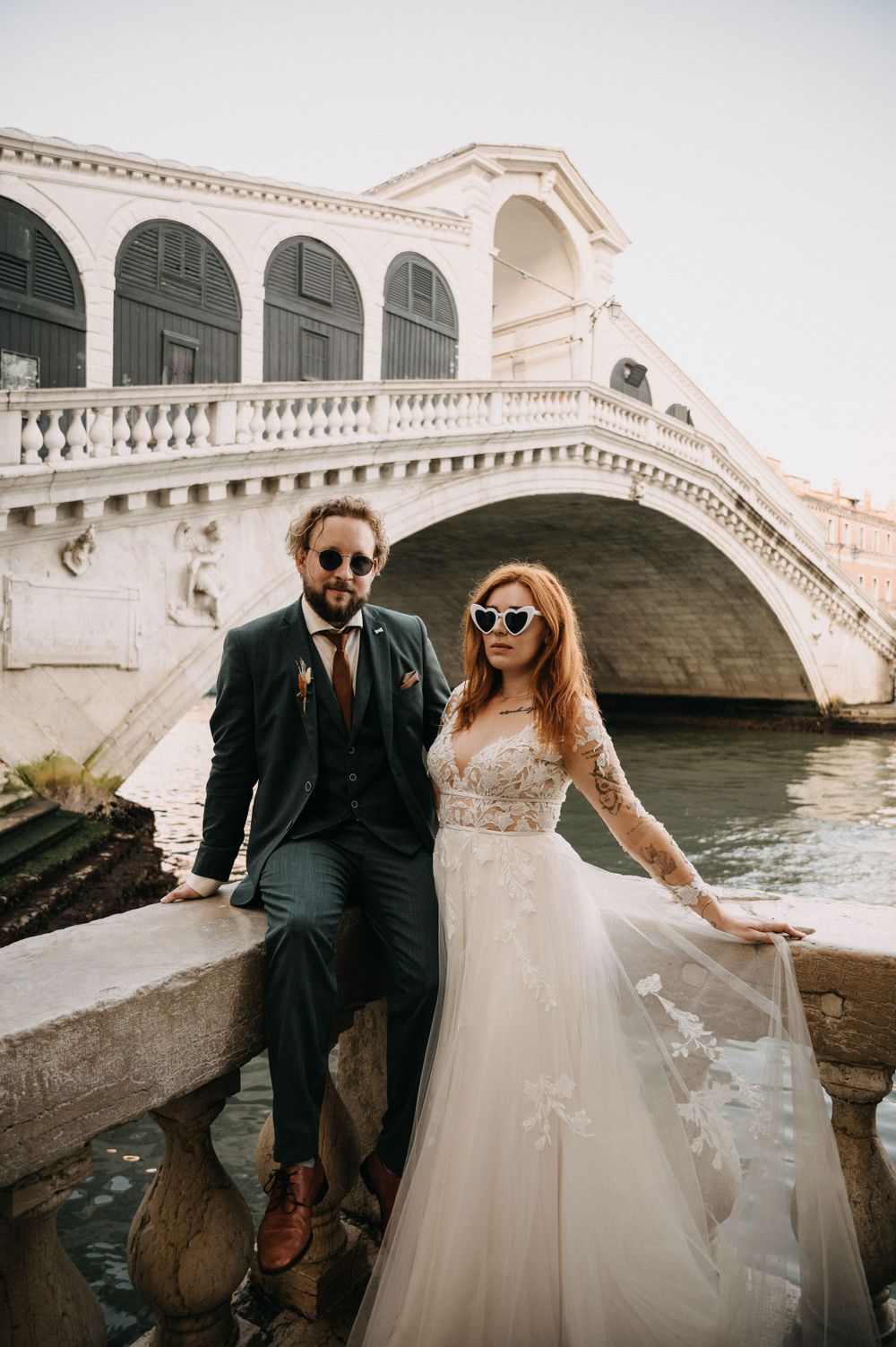 Stylish bride and groom pose at the iconic Rialto Bridge in Venice wearing sunglasses.